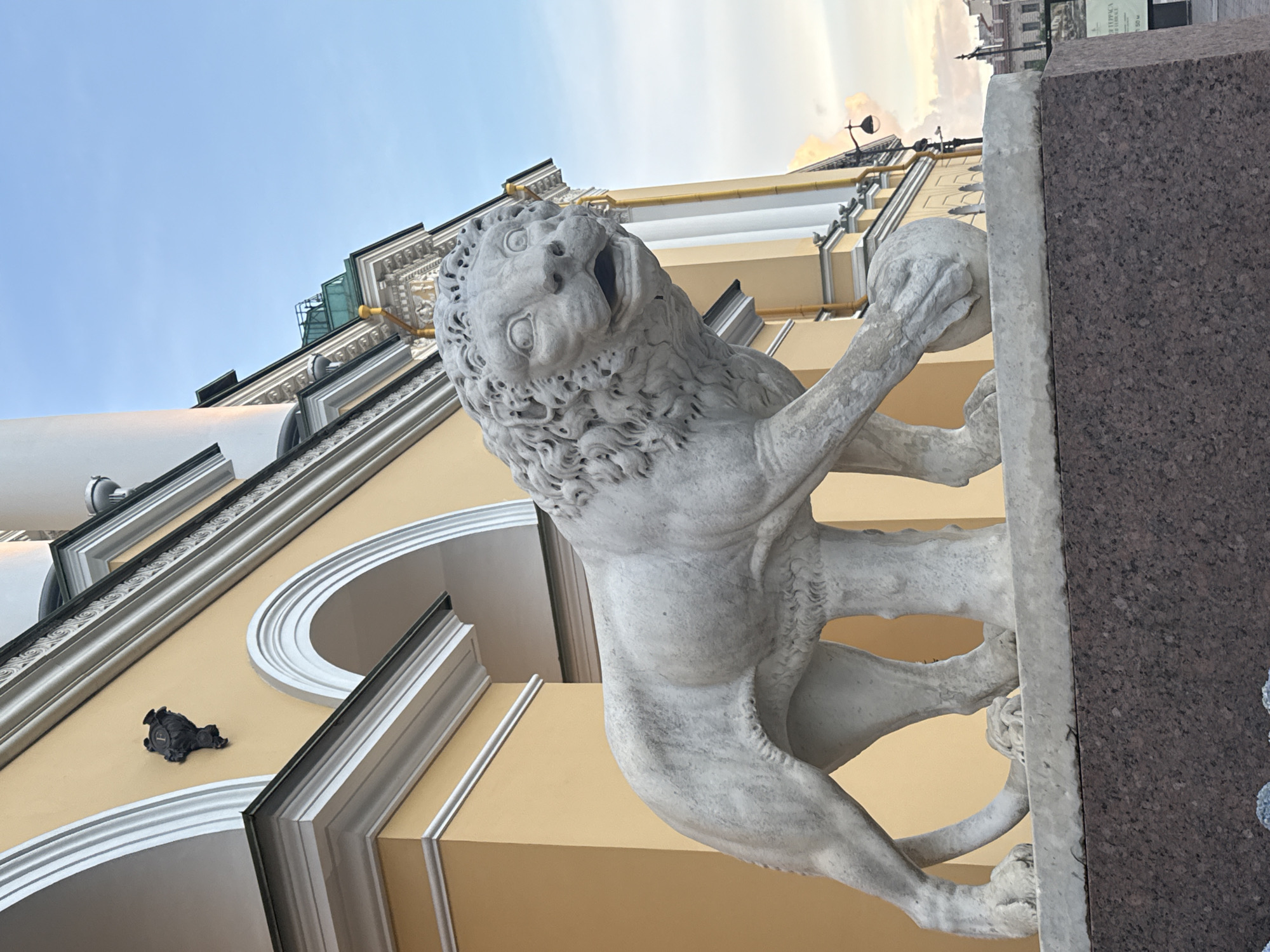 A stone statue of a lion with its paw resting on a ball stands in front of a yellow, neoclassical building. The lion is depicted with its mouth open and eyes wide, as if in mid-roar. The building has arched windows and decorative moldings. The sky is a soft blue with some clouds. The scene appears to be in Saint Petersburg, Russia, likely at the Lion Bridge, which features similar lion statues guarding its embankments.