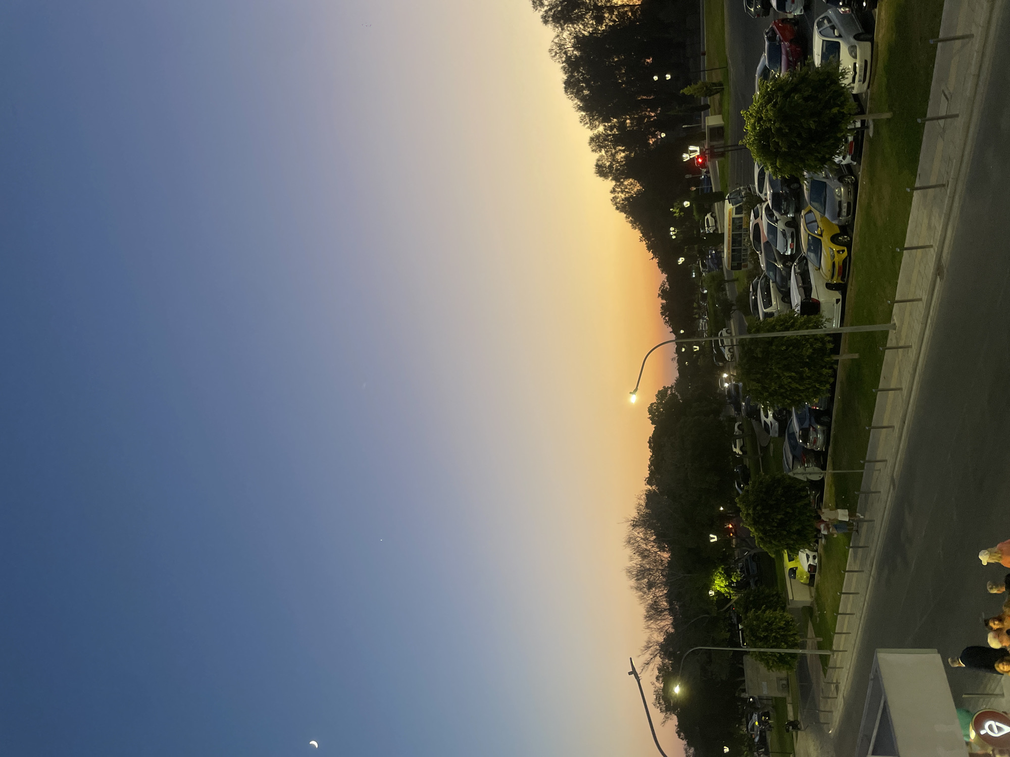At dusk, a crescent moon and a star are visible in the clear blue sky, which transitions to a soft orange and yellow gradient near the horizon. A line of trees, some bare and some with foliage, forms a dark silhouette against the colorful sky. Below the trees, a parking lot is filled with numerous cars of various makes and colors, including a prominent yellow car and a white sports car. Streetlights are illuminated, casting pools of light on the pavement and parking lot. Two people stand in the parking lot near the yellow car, appearing to be in conversation. Further down, a road is visible with parked cars on the side and a few people walking on the sidewalk. In the foreground, a group of people are gathered, some standing and some sitting. The overall scene depicts a parking area and roadside in Paphos Municipality, Cyprus, during the transition from day to night.