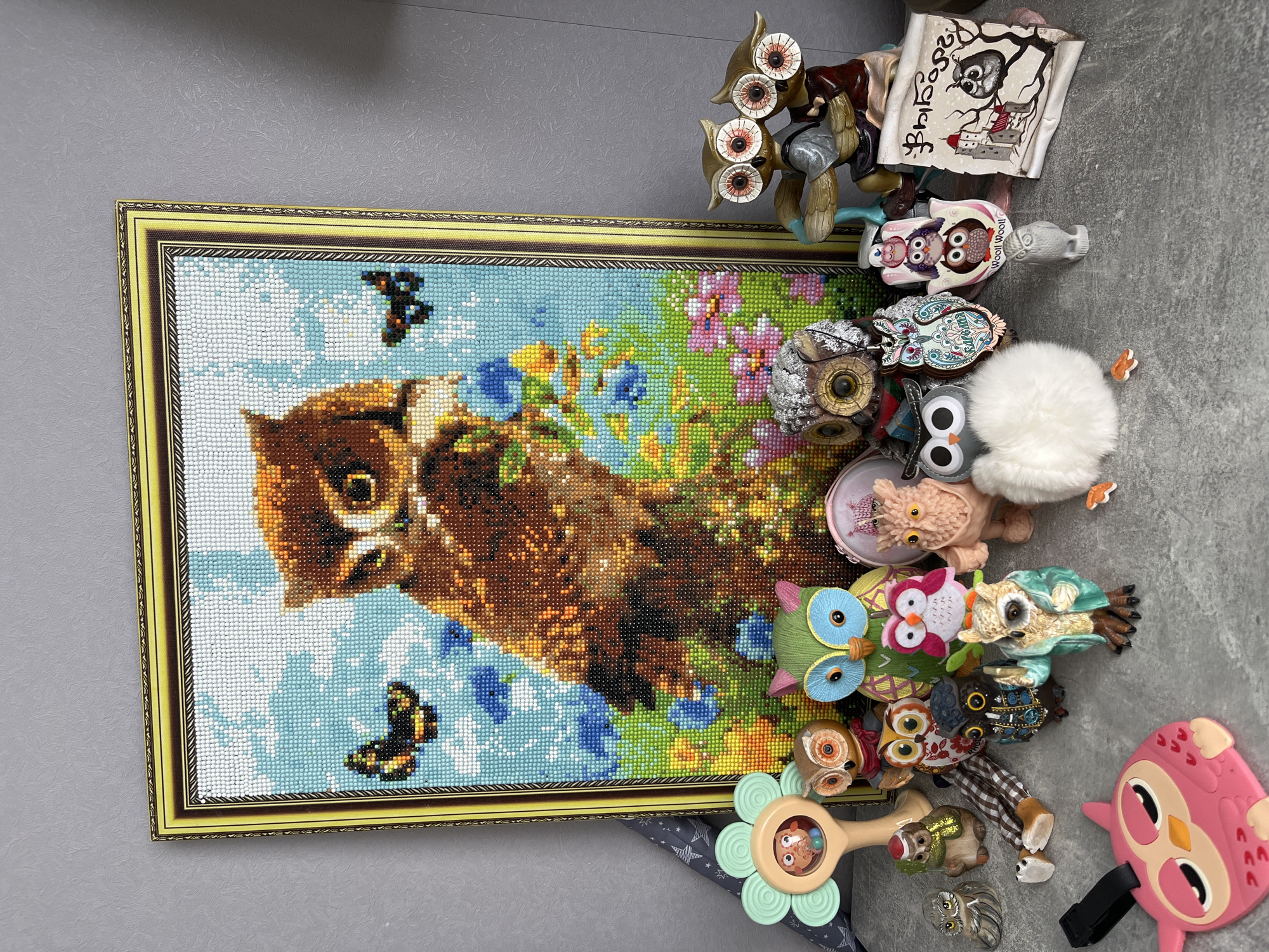 The image shows a collection of owl-themed items. In the center is a framed, diamond-painted artwork of an owl with butterflies and floral background. Adjacent to the artwork are various owl figurines and toys, including a multi-eyed metal sculpture, a scroll with an owl illustration, ceramic and plush owls, and a child's rattle with an owl design. The items are arranged on a gray surface, possibly a floor or table. There is no information to suggest the picture was taken in the city of Shushary, Russia.