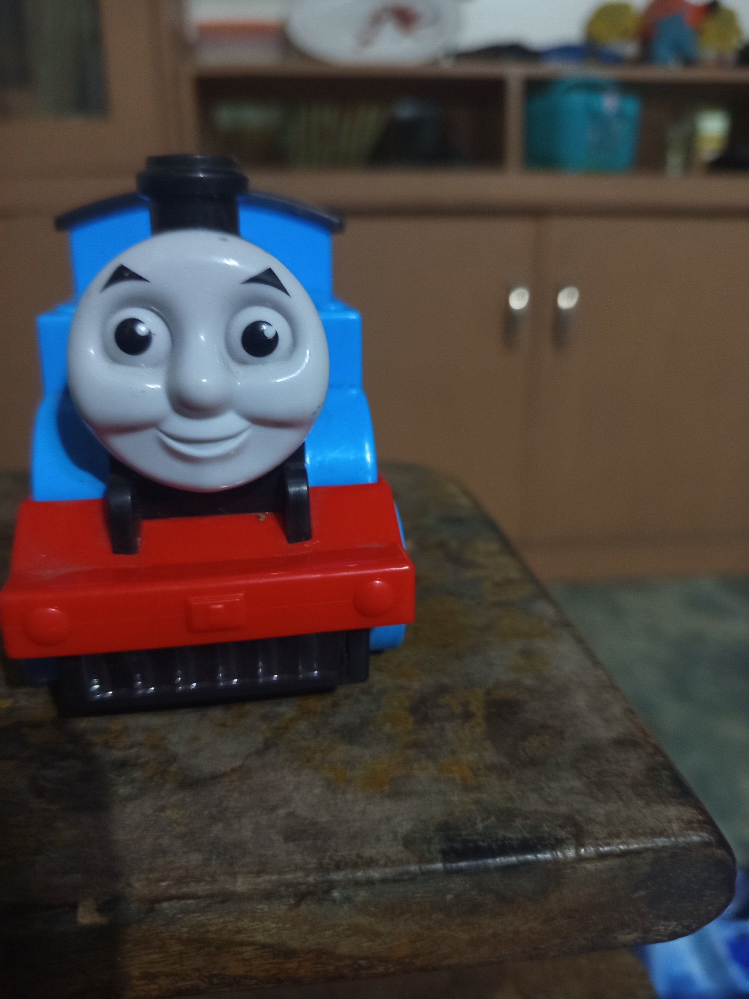 A close-up, eye-level shot features a toy Thomas the Tank Engine positioned on a textured wooden surface. The toy train is predominantly blue and red, with a white face characterized by large, black eyes and a wide smile. The background is blurred but appears to show a wooden cabinet with two handles, suggesting an indoor environment. The overall lighting is moderate, casting faint shadows. The image lacks any identifiable landmarks or context to place it within the city of Ngablak, Indonesia.