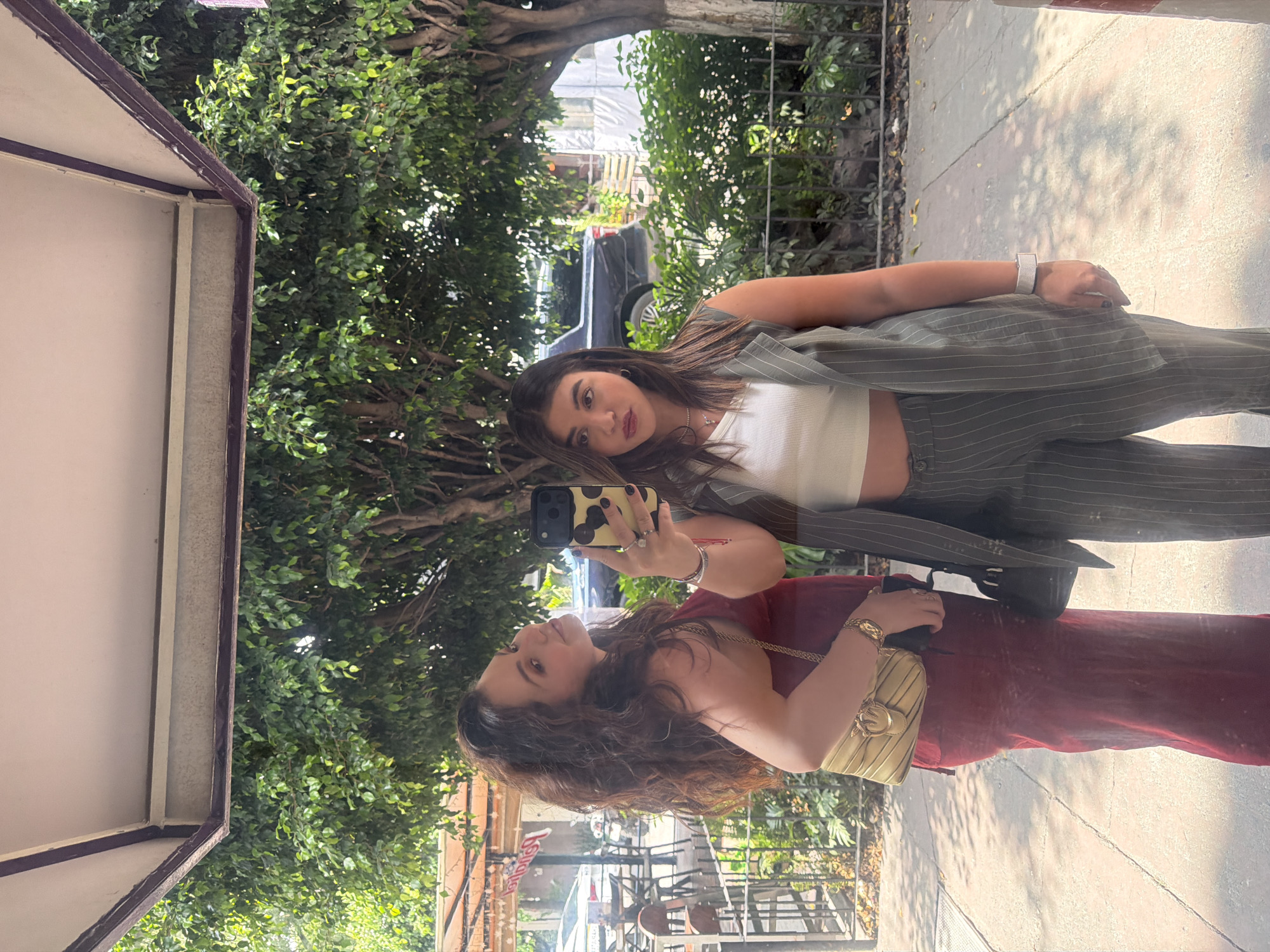 A photograph shows two women in the reflection of a pane of glass, likely a store window. The woman in the foreground is wearing a maroon top and carrying a gold handbag. She is holding a cell phone with a cow-print case, apparently taking a selfie. Behind her, the woman is wearing a white crop top and a pinstripe suit. They are standing on a sidewalk beside a street with trees. Beyond the street is a building with tables and chairs suggesting a restaurant or cafe. The location is most likely in Mexico City, Mexico.