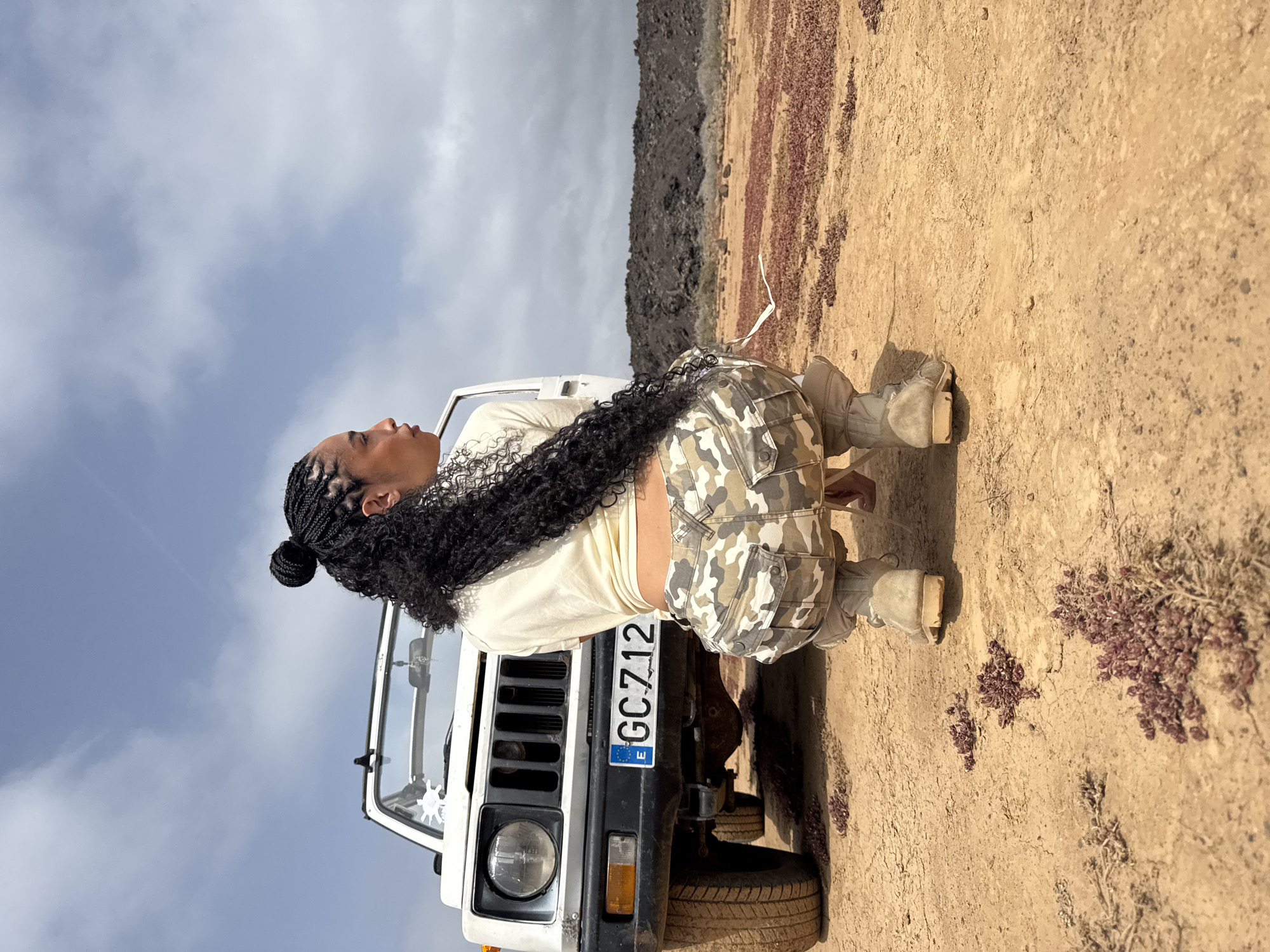 A woman is squatting behind the front of a white vehicle with Spanish license plate "GC712". The vehicle appears to be a 4x4. The woman has dark hair braided and pulled into a bun, with some of the hair hanging down her back. She is wearing a light-colored top, camouflage-patterned shorts, and light tan boots. The background is a dry, sandy landscape. In the distance, there is a hill. The sky is visible and is cloudy. The location appears to be in the city of Tinajo, Spain.