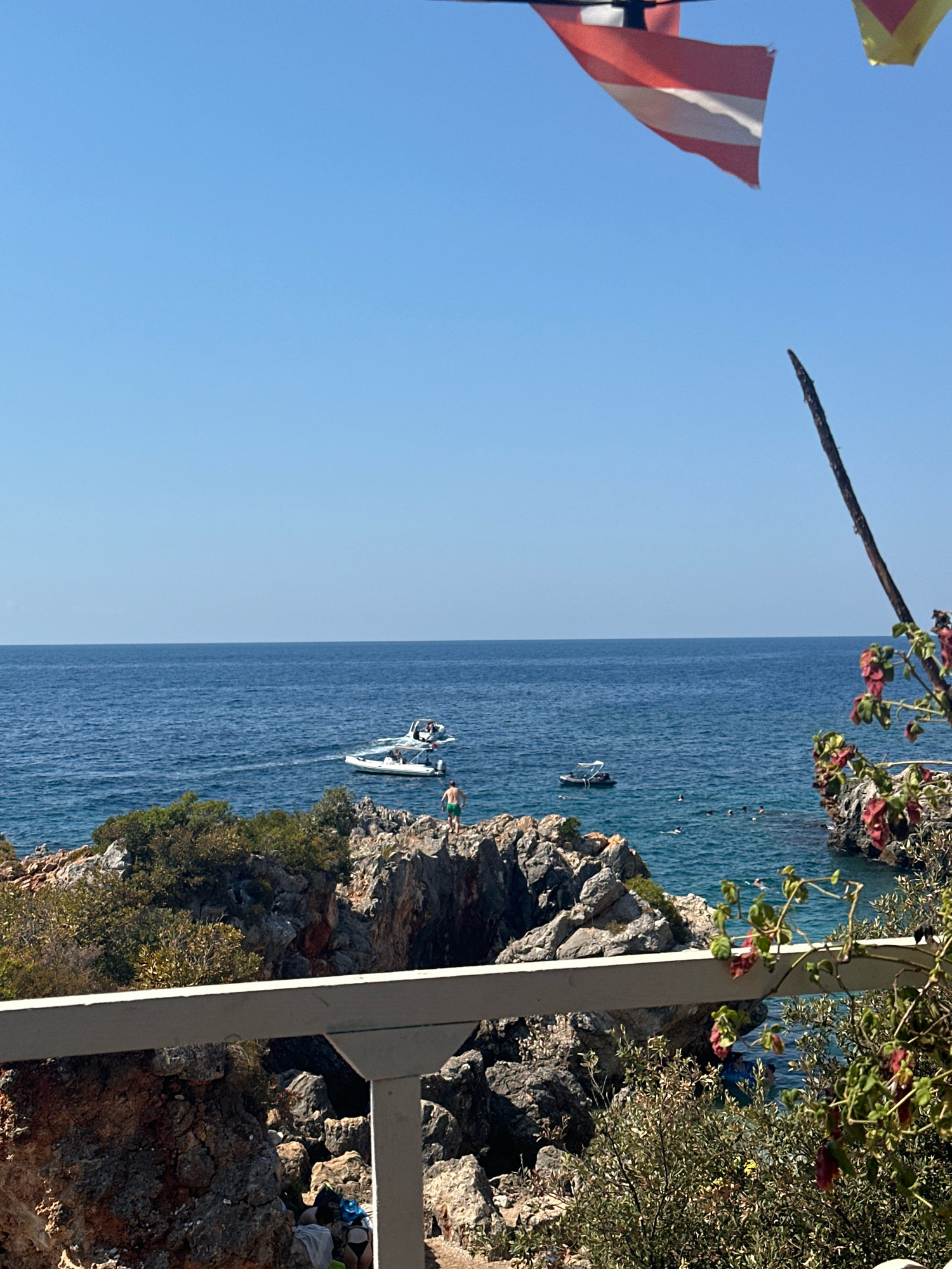 The image depicts a rocky coastline on a clear, sunny day. The deep blue sea stretches to the horizon under a bright blue sky. In the foreground, a white railing runs horizontally across the lower portion of the image, with lush green foliage and some reddish flowers growing on the right side.
The rocky shore is covered in various shades of brown and gray, with patches of green vegetation clinging to the rocks. There are several boats in the water. One white boat, possibly a speedboat, is near the shore, leaving a wake behind it. Another smaller, darker boat is further out. Several people are visible in the water, swimming and enjoying the sea. One person in green shorts stands on a rocky outcrop on the right side of the image.
In the upper right corner, parts of two flags are visible, suggesting a nautical or recreational area. The overall scene conveys a sense of a peaceful summer day by the sea, with people engaged in various water activities. The specific location in the city of Unknown, Unknown is not determinable from the visual information.