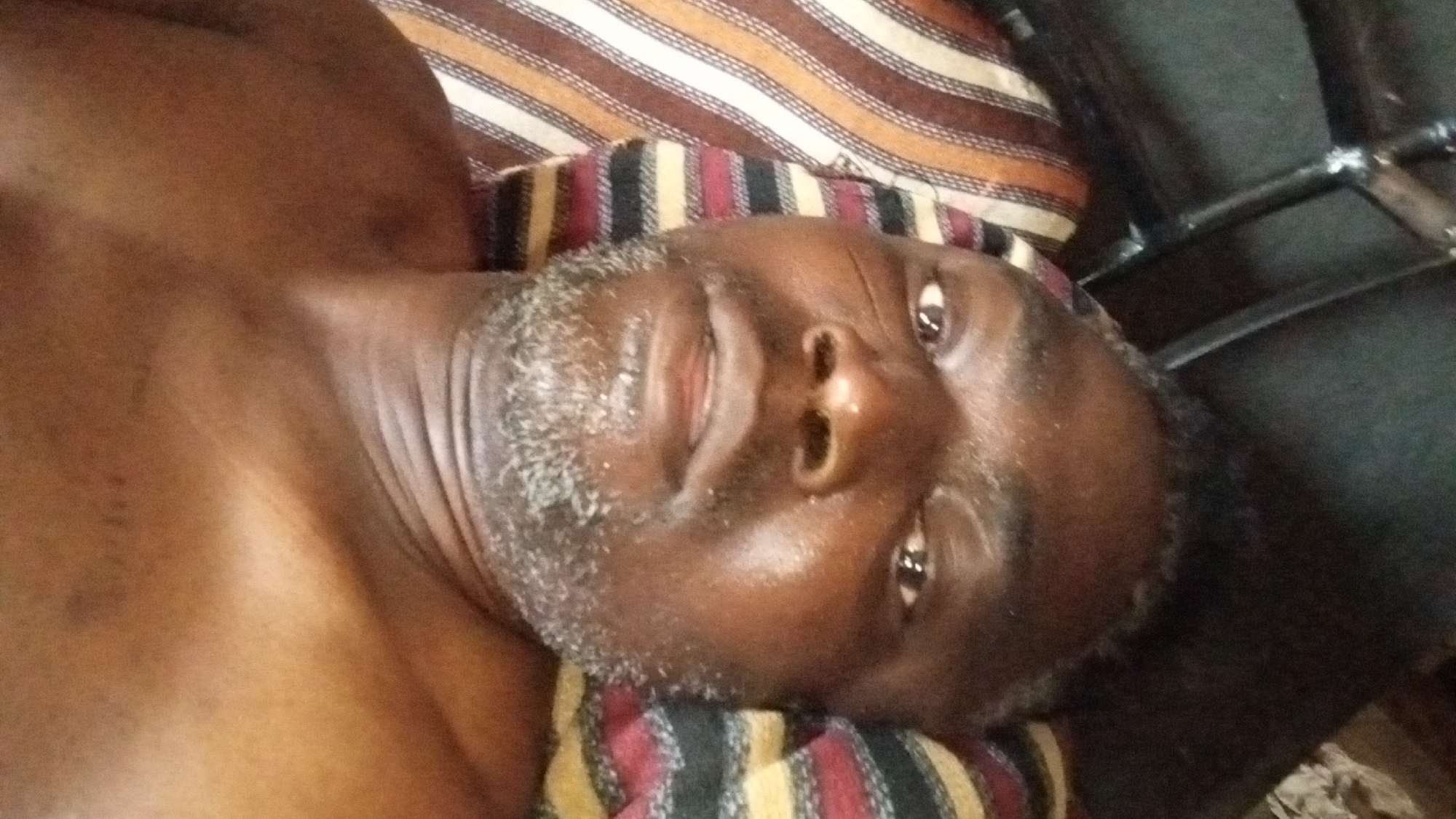The media file displays a close-up of a man with dark skin, positioned with his head towards the bottom left of the frame and his body extending towards the top right. He is lying down, looking directly upwards at the camera with a calm expression and a slight smile or smirk. He has a short, patchy beard that is predominantly grey or white. His head rests on a pillow or blanket with distinct horizontal stripes in shades of brown, beige, red, and black. To the right side of his head, a dark, metallic structure, possibly part of a bed frame or chair, is visible. The scene is indoors, characterized by somewhat dim lighting. No discernible text is present in the image.
