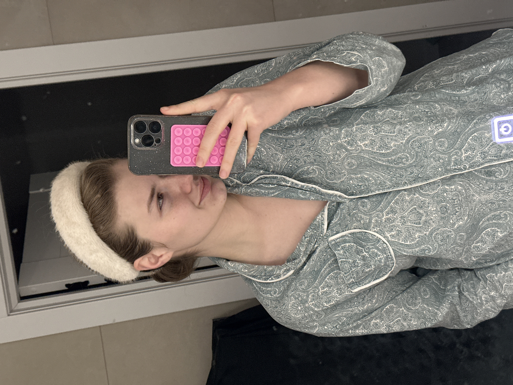 A woman is taking a selfie in a mirror. She is wearing a paisley-patterned pajama set with white piping and a fluffy white headband. She is holding a dark-colored smartphone with a pink pop socket in her right hand. The background shows a tiled floor, a dark-colored object that may be furniture, and a mirror framed in a white frame. There is no indication in the image that the location is in the city of Kazan, Russia.