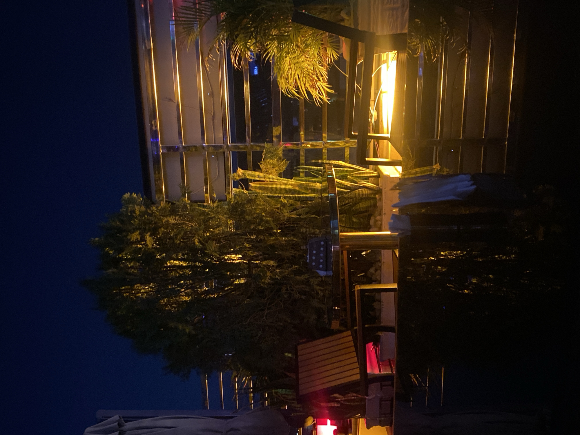 A nighttime outdoor scene in Sabon Gari West, Nigeria, features a contemporary patio setting. A tall, leafy green tree illuminated by warm yellow lights stands prominently in the center. To its left, a portion of a light-colored umbrella or canopy is visible. In front of the tree, a wooden chair with vertical slats is positioned near a round glass-top table with a reflective surface and metallic cylindrical legs. The table appears to have a grill or a similar dark object on it. Surrounding the table and chair are planters with various plants, including what appear to be tall, spiky cacti or succulents. Behind these elements, a sleek, modern structure with horizontal metallic slats and integrated warm yellow lighting creates a backdrop. To the right, a palm-like plant with fronds catches the light. The ground in the foreground is dark and appears wet or reflective, possibly indicating a pool or polished surface. The overall atmosphere is one of relaxed, illuminated outdoor dining or lounging.