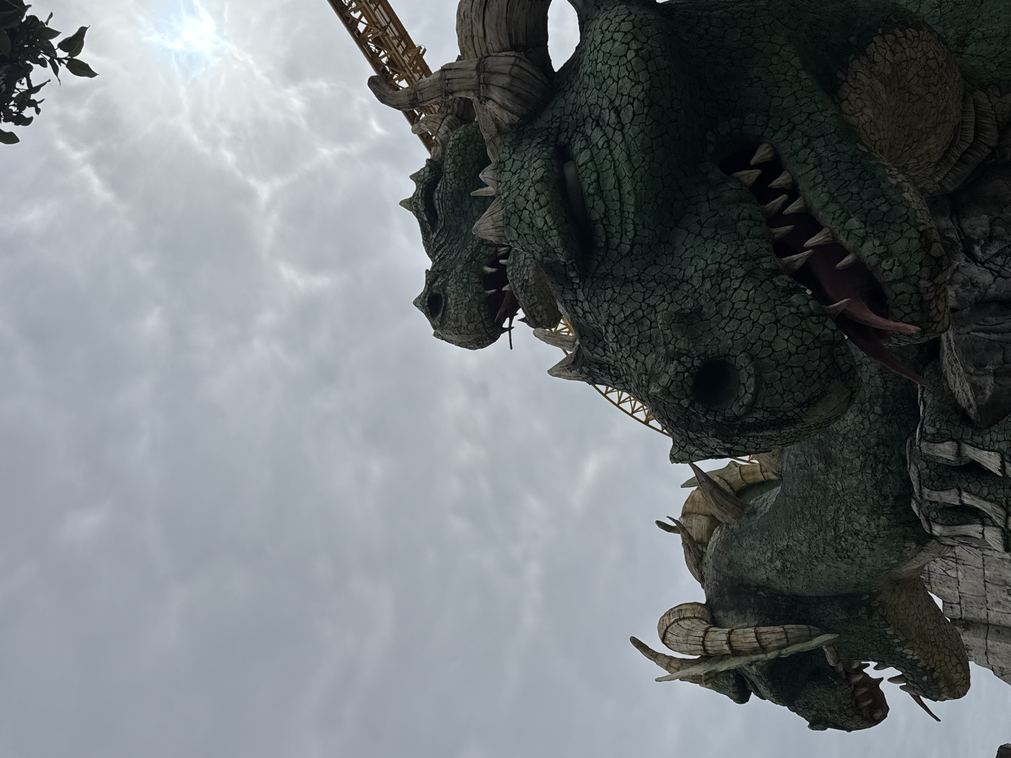 The image displays a low-angle perspective of large, dark green dragon statues integrated into a themed ride structure. In the foreground, a dragon's head is prominently featured, facing right with its mouth agape, revealing multiple sharp white teeth and a pink tongue. Its textured skin is characterized by intricate, dark green scales. Behind and to the left of this main figure, additional sections of dragon heads and bodies are visible, similarly scaled and adorned with horn-like protrusions, all appearing to form a unified, rocky facade. A yellow, latticed roller coaster track is observed curving across the upper right background, passing behind the dragon elements. The sky above is heavily overcast with grey clouds, exhibiting a diffused bright spot in the upper right quadrant, indicative of the sun attempting to penetrate the cloud cover. A small cluster of dark green foliage is partially visible at the extreme top right edge. No human figures or specific actions are depicted. This scene is identified as part of the "Dragon Roller Coaster" (also known as "Knyaz Vladimir") attraction located at Sochi Park in the Adler district of Sochi, Russia.