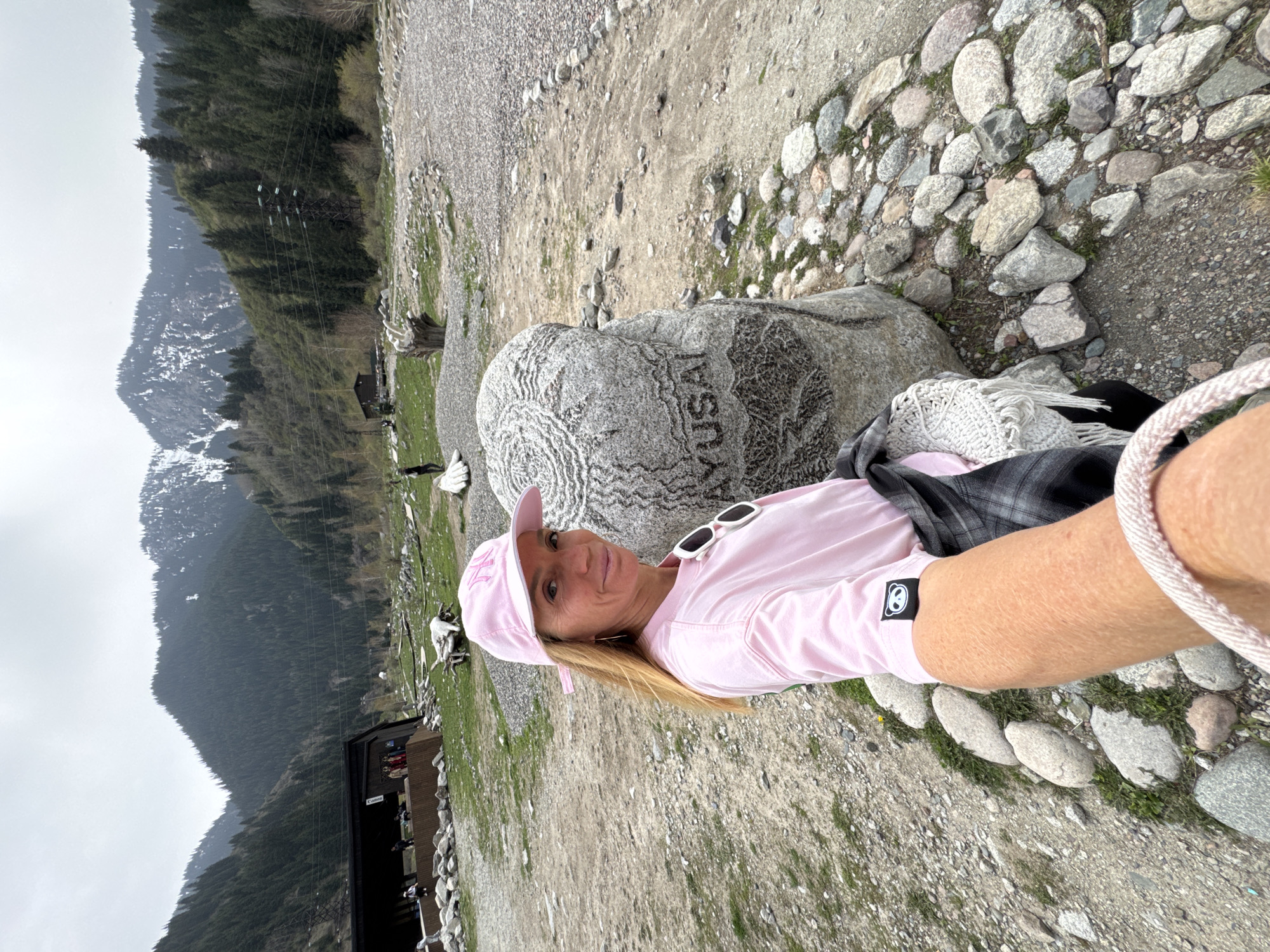 A woman is captured in a selfie, posing outdoors in a mountainous, rocky landscape. She is in the foreground, smiling at the camera, wearing a pink baseball cap, white sunglasses perched on her cap, and a light pink long-sleeved shirt with a black and white patch on the sleeve. A black and white plaid shirt is tied around her waist, and a white knitted scarf or blanket rests on her shoulder.
To her right, a large gray stone is prominent, with the word "AYUSAI" carved into its surface. The ground around her is composed of dirt, gravel, and numerous small and large rocks, with patches of green grass interspersed.
The background features towering mountains covered in dark green coniferous forests. Some mountain peaks show remnants of snow, suggesting a higher elevation or cooler climate. In the middle distance, a dark building, labeled "Canteen," is visible, with small animal figures (possibly goats or sheep) nearby. A large metal power pylon and overhead electrical lines stretch across the scene, indicating human infrastructure within the natural environment.
The sky is overcast, suggesting a cloudy day, but it is clearly daytime. The woman appears content and happy, enjoying her outdoor excursion. The text "AYUSAI" on the stone is a significant location marker, aligning with the provided context of Almaty, Kazakhstan, where Ayusai is a known recreational area.