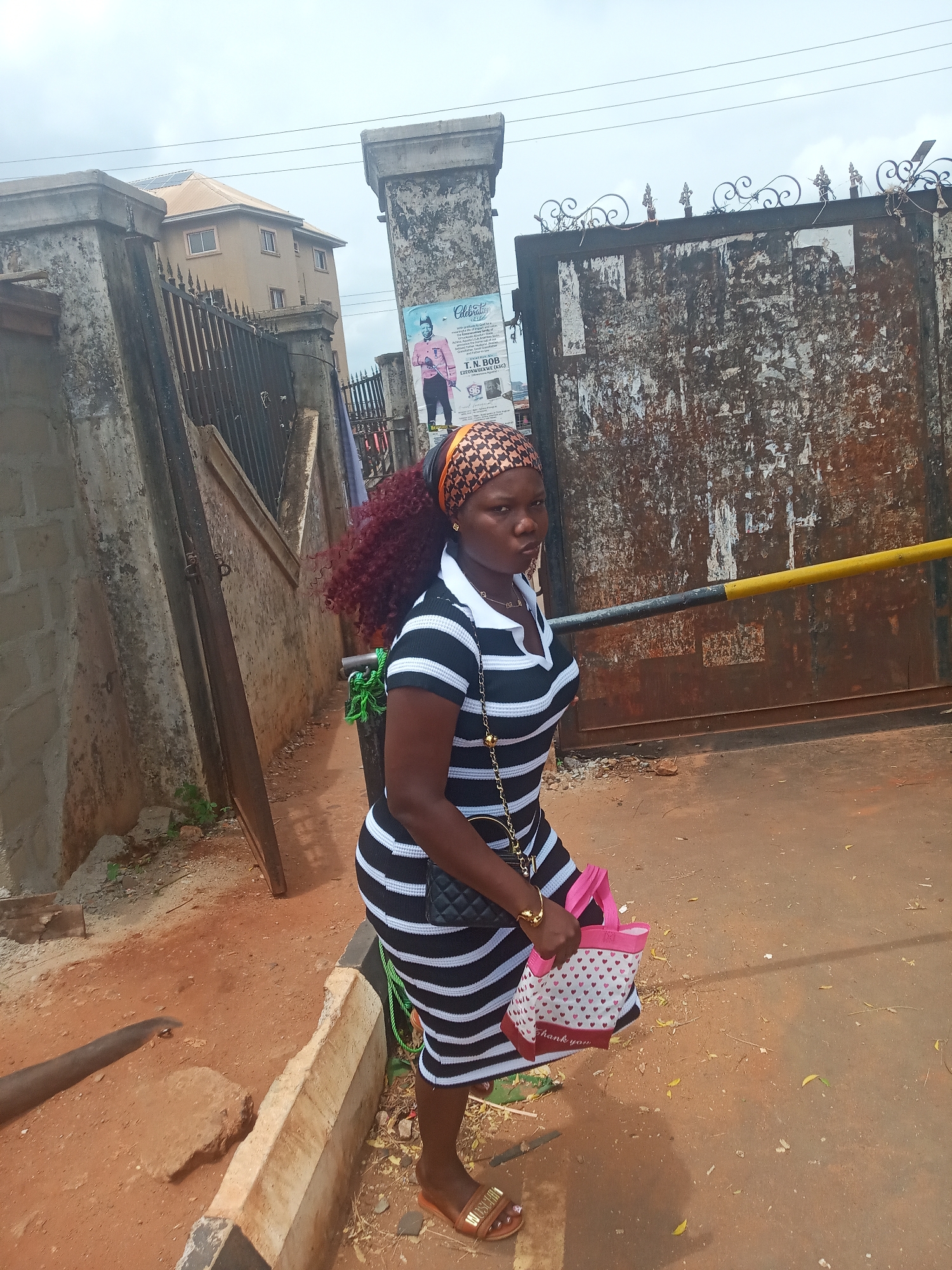 A full-body photograph shows a woman standing outdoors in what appears to be an urban or semi-urban setting. She is positioned slightly to the left, looking directly at the camera with a neutral expression.
The woman has dark skin and is wearing a black and white horizontally striped dress or top with a white collar. Her head is adorned with a patterned headscarf in shades of brown, orange, and black, from which long, curly, reddish-brown hair flows. She wears a gold chain necklace and gold earrings. A black quilted shoulder bag with a gold chain strap hangs from her left shoulder, and in her left hand, she holds a pink and white tote bag decorated with red hearts and the words "thank you." She completes her attire with brown slide sandals, one of which partially displays "MOSCHINO."
The background features a sturdy concrete pillar to the left, and a large, heavily weathered and rusty metal gate with decorative elements at the top to the right. This gate is also flanked by a concrete pillar, upon which a partially visible poster with the word "Celebrati..." and the name "T.N. BOB" can be seen, depicting a man in a pink suit. A black and yellow striped barrier bar crosses horizontally in front of the gate. In the distance, a light brown multi-story building with numerous windows is visible under a somewhat overcast sky. The ground is unpaved, consisting of reddish-brown dirt, with a low concrete curb running parallel to the woman. The scene is illuminated by daylight.