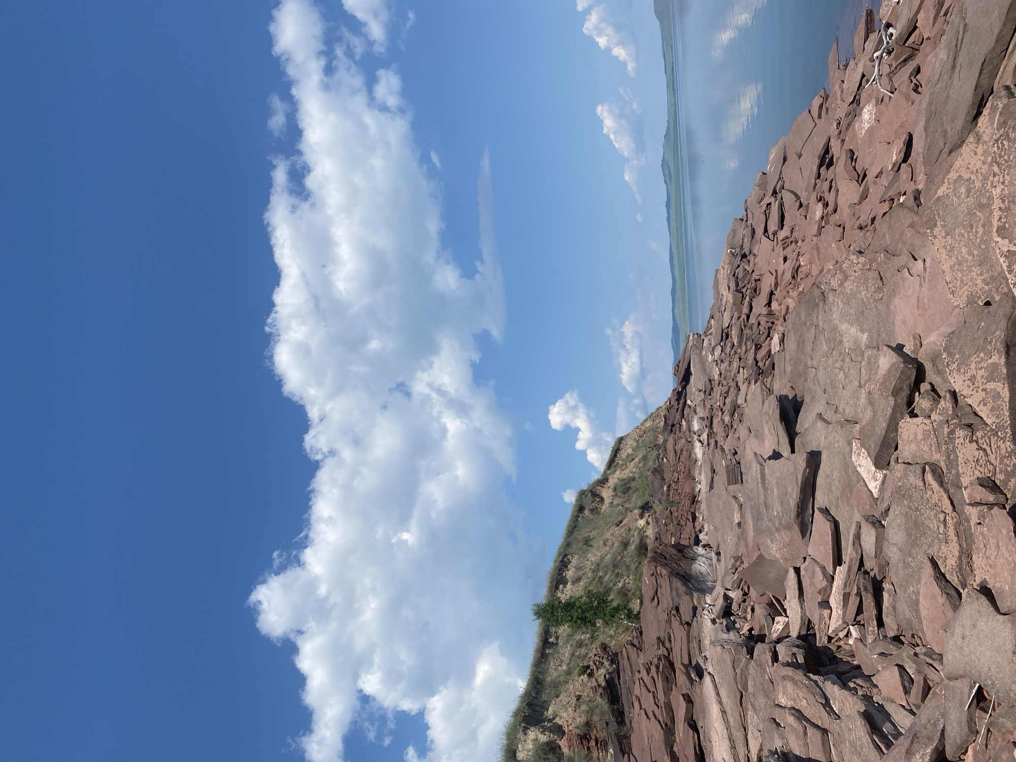 This is a photograph taken from the shore of a body of water in Ust'-Erba, Russia. In the foreground, the shore is covered with numerous red, flat rocks of varying sizes. A grassy, eroded cliff rises to the left of the rocky shore. A single tree stands on the cliff face. The body of water in the midground is calm and reflects the sky. In the background, a distant shoreline with rolling green hills is visible. The sky is a bright blue with scattered, fluffy white clouds. There are no people or animals visible in the image.