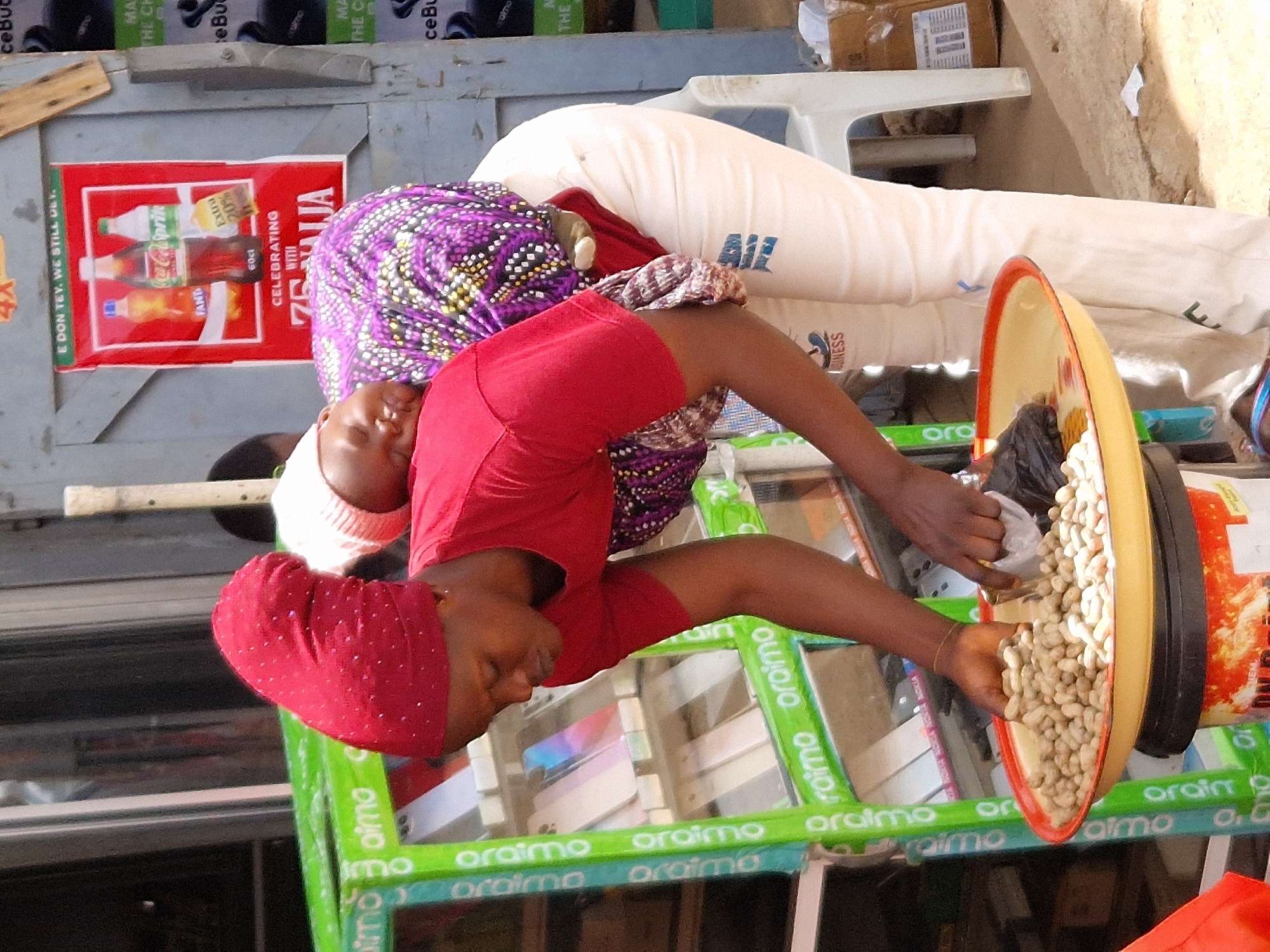 In an outdoor urban environment, a woman wearing a red headscarf and a red shirt is seen from the chest down. She is tending to a round, yellow tray filled with peanuts, which rests on top of a colorful container. Her hands are actively engaged with the peanuts, with one hand scooping them into a plastic bag, and the other hand partially submerged in the peanuts. Behind her, a baby wrapped in a purple and white patterned cloth is being carried on her back. The baby is wearing a white cap. To the left, a green display stand with multiple shelves is visible, showcasing various items, possibly phone cases or accessories. A poster advertising drinks, including Coca-Cola, Sprite, and Fanta, is displayed on a wall behind the woman and baby. The ground appears to be dirt or sand, and a white plastic chair is situated to the right of the frame.