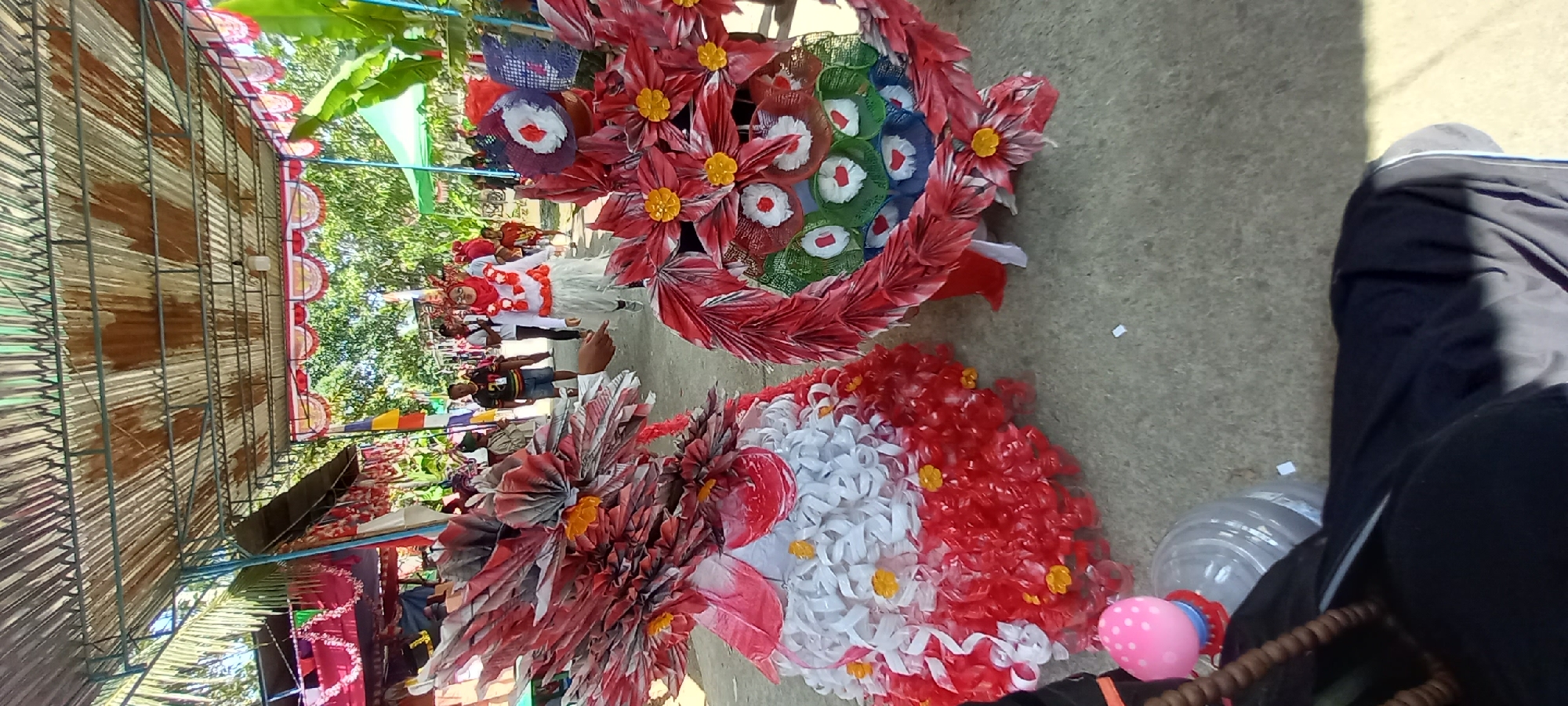 In Sidamulya, Indonesia, two elaborate costumes, one resembling a tiered skirt and the other a circular headdress, are displayed outdoors under a corrugated metal roof. The costumes are constructed from red and white recycled materials, likely plastic bottles, folded into decorative elements resembling petals and feathers, with yellow accents. A person in a white outfit and a red headscarf walks behind the costumes. In the background, other individuals are seen walking along a paved pathway, with palm trees and other foliage visible beyond the walkway. The scene appears to be part of a local celebration or parade.