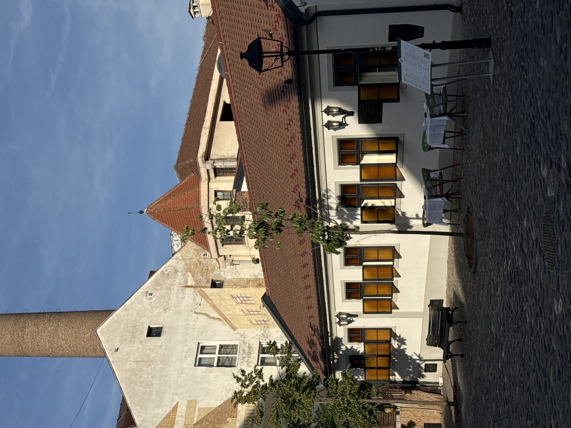 The image depicts a street scene in Belgrade, Serbia. The foreground shows a cobblestone street with a bench on the right side. Tables and chairs with white tablecloths are set up next to a white building with black-framed windows, some of which have amber-colored glass. A lamp post and a menu stand are near the tables. To the left, other buildings with red-tiled roofs are visible. In the background, a tall brick chimney rises against a blue sky. The overall atmosphere is bright and sunny.
