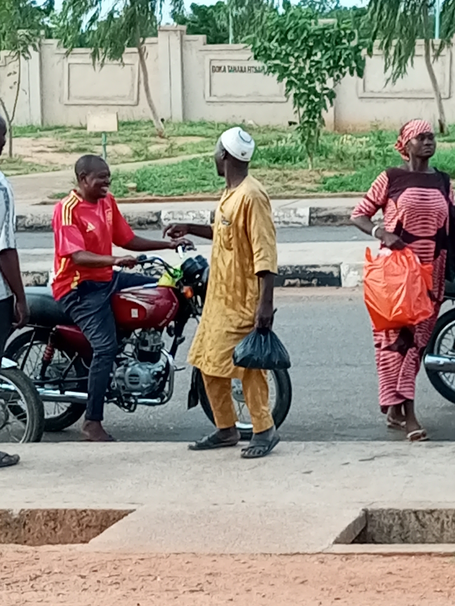 In Gombe, Nigeria, three individuals are depicted outdoors in daylight, standing near a red motorcycle parked on a paved surface. On the left, a man in a red and yellow t-shirt and dark trousers is seated on the motorcycle, smiling and looking to his right. To his right, another man in a yellow tunic and trousers faces the seated man, holding a black plastic bag in his left hand. A woman stands further to the right, wearing a pink and black patterned dress and headscarf, and holding an orange plastic bag in her right hand. In the background, a concrete wall with decorative panels and trees with green foliage are visible. The ground is a mix of paved areas and dirt.
