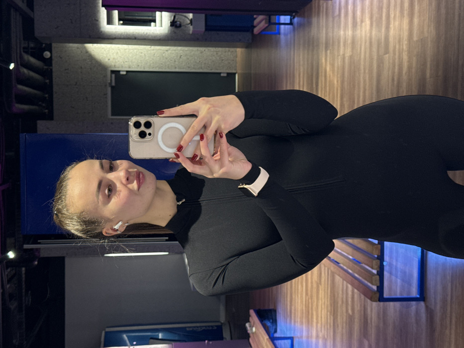 A young woman in a black athletic jumpsuit takes a selfie in a gym. She holds a smartphone with a clear MagSafe case. Her nails are painted red. She wears a smart watch on her left wrist and an earbud in her left ear. The gym has wood floors, blue accents, and exercise equipment. A mirror reflects a blue structure.