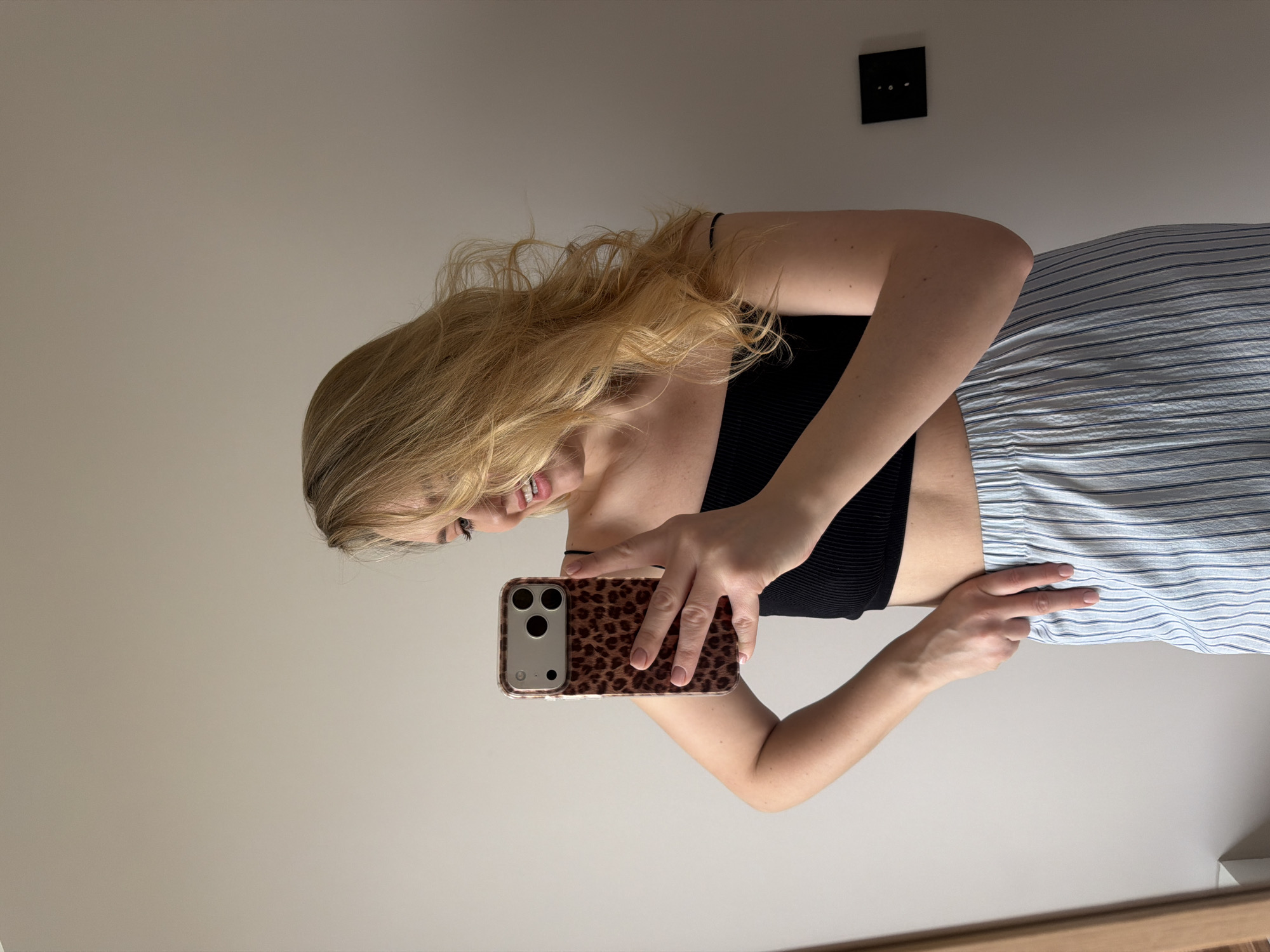 A young woman is captured in a mirror selfie, originally oriented 90 degrees counter-clockwise. She has long, wavy blonde hair and a cheerful expression, indicated by her broad smile. She is wearing a black, ribbed crop top or bra and light blue and white vertical striped pleated pants or a skirt. Her right hand holds an iPhone, which has a distinctive brown and black leopard print case, directed towards the mirror to capture the reflection. Her left hand rests on her hip, forming a peace sign gesture with her index and middle fingers. The background is a plain, light-colored wall. A black square electrical outlet or switch is visible on the wall in the upper right portion of the image. The lighting suggests it is daytime. There is no visible text in the image. The location context provided is Dreiliņi, Latvia.