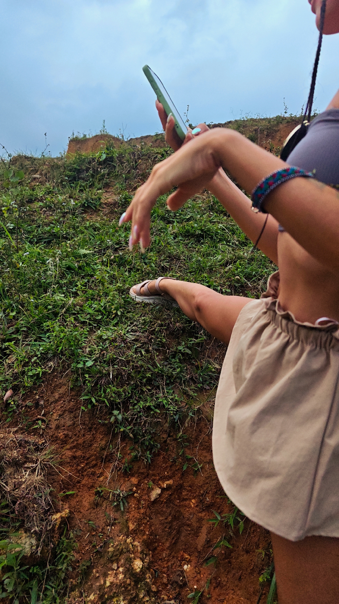 A high-angle shot captures a person sitting on a verdant hillside, characteristic of Nậm Dịch Commune, Vietnam. The individual, partially framed from the waist up, is engaged with a light green smartphone held in their right hand. Their hands feature long, light-colored polished nails. A colorful, woven bracelet adorns their left wrist. The person wears light-colored, ruffled shorts and a dark-colored top, with one leg extended, revealing a light-toned sandal. The foreground and midground are dominated by lush green grass, various plants, and exposed reddish-brown soil, suggesting a natural, earthy terrain. The background features a gentle rise of the hillside against an overcast, pale sky, indicating a cloudy day without direct sunlight. The scene depicts a moment of modern technological interaction within an unadorned, natural outdoor setting. No discernible text is visible within the image.