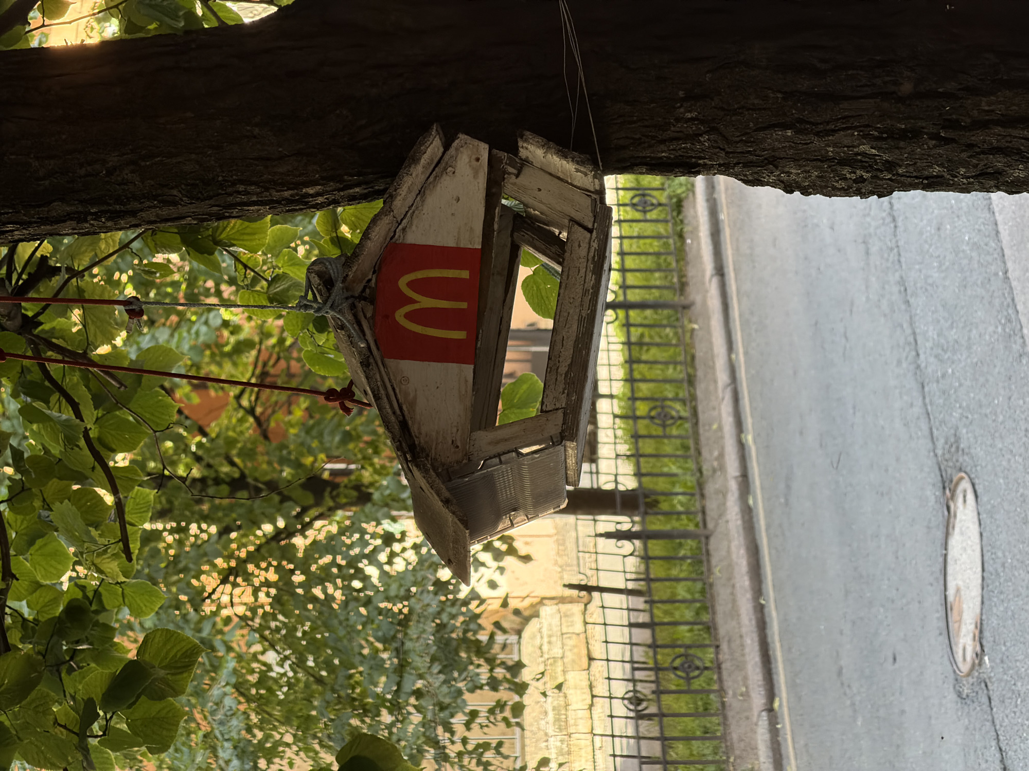 A rustic, white wooden birdhouse with a red rectangle featuring a yellow McDonald's "M" logo is hanging from a tree in Saint Petersburg, Russia. The birdhouse is affixed to a thick tree trunk with white twine. Behind the tree and birdhouse, a metal fence with decorative elements encloses a green grassy area. Beyond the fence, a light-colored building with arched windows is visible. A paved road with yellow lane markings runs in front of the fence, and a circular manhole cover is present in the road. The scene is illuminated by dappled sunlight filtering through green leaves.