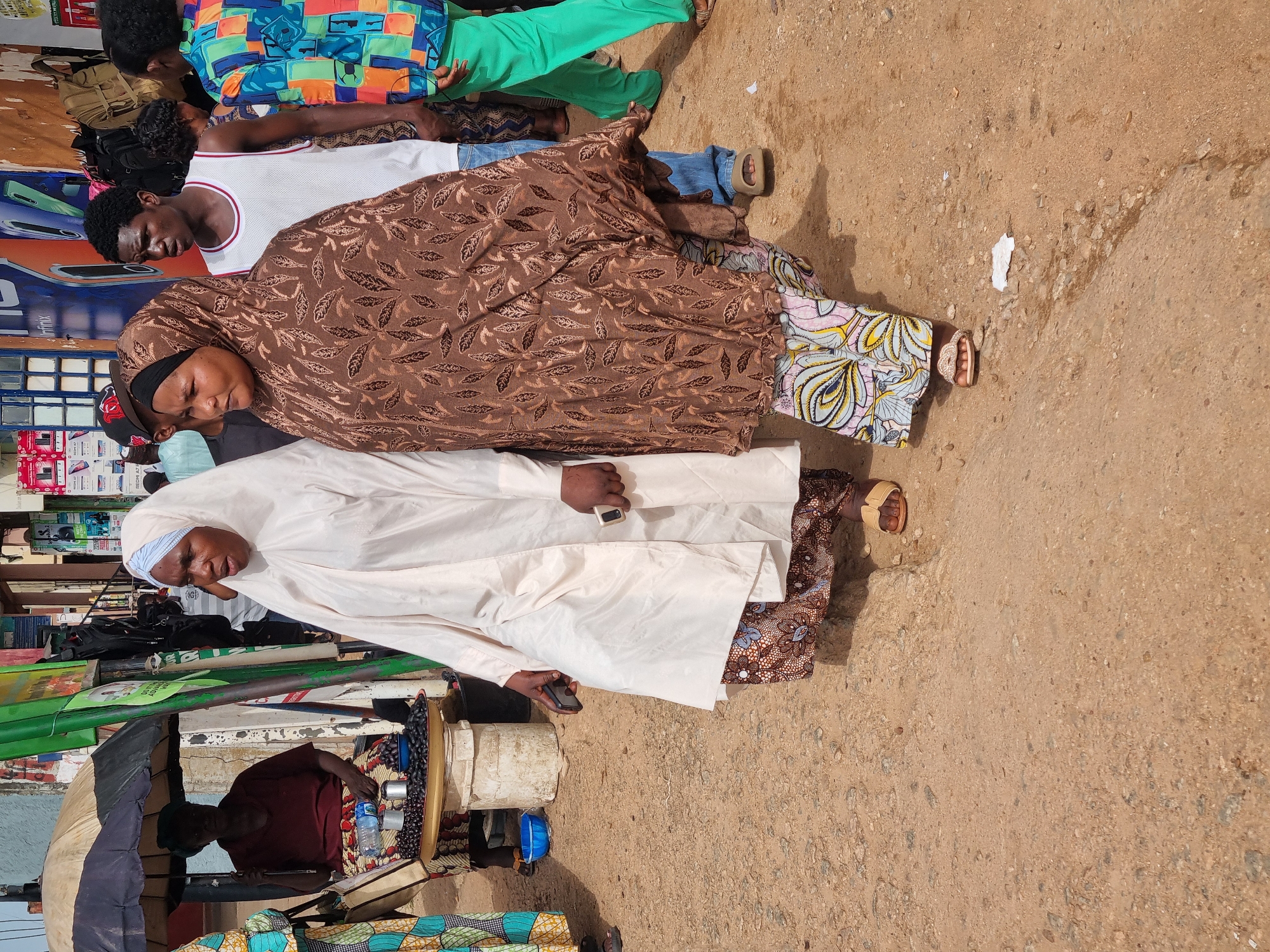 Two individuals are positioned in the foreground of the image, standing on a dusty, uneven ground. The person on the left is wearing a white robe and a light-colored headscarf, with their eyes closed and head tilted back, suggesting they may be resting or praying. They are holding a dark-colored mobile phone in their right hand. To their right, another individual is seen partially, wearing a brown patterned headscarf and a long brown garment with a leaf-like pattern. Their eyes are also closed, and their head is slightly bowed. This person is wearing sandals.
Behind them, other individuals are visible. One person is wearing a white tank top with red trim and appears to be looking down. Another individual is dressed in a brightly colored, geometric-patterned shirt and green pants. The scene appears to be outdoors, with a sandy or dirt ground. In the background, there are glimpses of what look like market stalls or shops, with various items displayed on shelves, including what seem to be mobile phones. There are also some green poles and signage.
The lighting suggests it is daytime, possibly mid-morning or afternoon. The weather appears to be warm and dry, consistent with the dusty environment. The overall impression is of a busy, potentially market-like setting in Jos, Nigeria, where people might be taking a break or engaged in quiet contemplation amidst the activity.