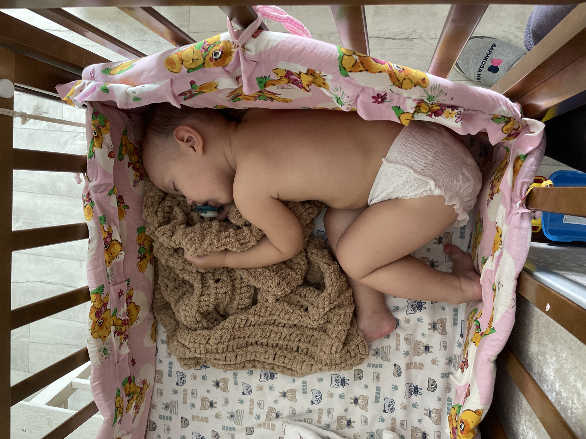 A baby is sleeping in a crib. The crib is lined with a pink bumper pad featuring cartoon animals. The baby is wearing a white diaper and has a pacifier in their mouth. A brown knitted blanket is near the baby. The crib sheet is patterned with bears. A label in Russian, "ВАРРИКОЗА НЕТ," is on an object out of the crib. The location is likely Perm, Russia, based on the Russian label.