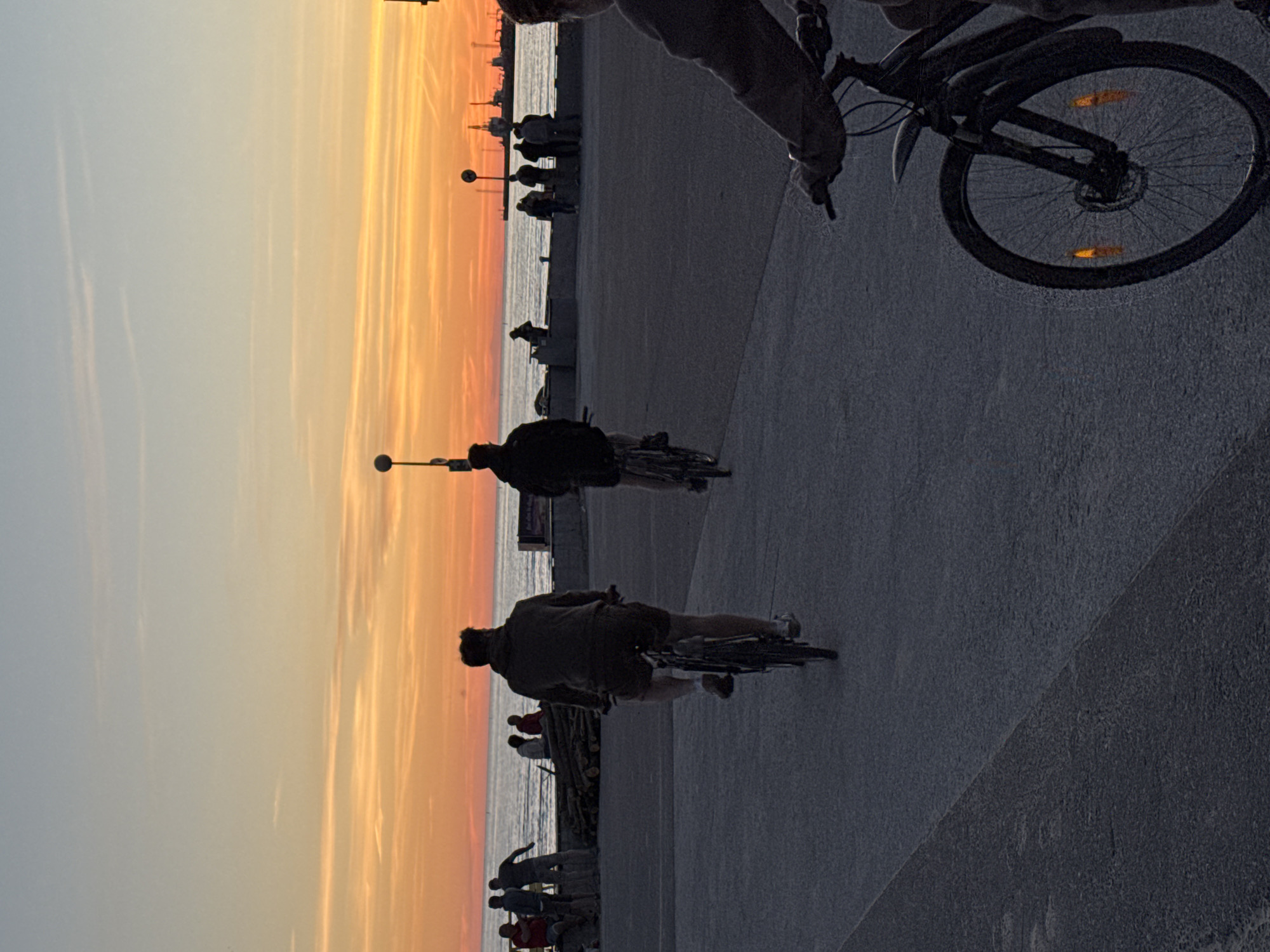 The image depicts a sunset over the sea, with a promenade in the foreground. Several individuals are seen cycling on the promenade, silhouetted against the vibrant orange and yellow hues of the sky. In the background, a group of people are gathered, and distant ships and industrial structures are visible on the horizon. The scene suggests a recreational activity taking place in an urban coastal setting, likely in the city of Tallinn, Estonia, given the architectural elements and maritime activity characteristic of the area. The overall mood is serene, with the natural beauty of the sunset contrasting with the urban environment.
