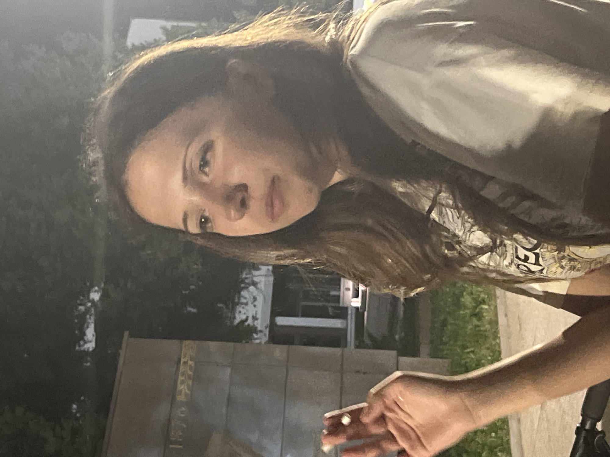 A young woman with brown hair is seen from the chest up, looking to the right. She is wearing a light-colored t-shirt with a graphic print. Her left hand is raised, holding what appears to be a cigarette. Behind her, a large stone structure with the inscription "1876" is visible. The background is dark, suggesting it is nighttime, with some trees and a building structure faintly discernible. The setting appears to be outdoors, possibly in a public space in the city of Vratsa, Bulgaria, given the historical marker.