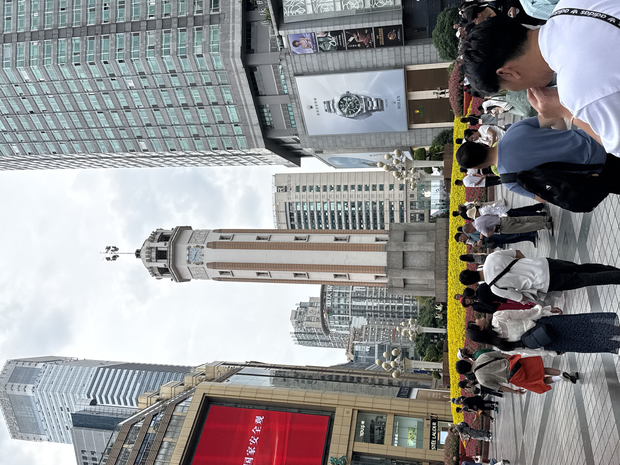 The image captures a bustling street scene in Yuzhong District, Chongqing, China, with a mix of architecture, commercial elements, and pedestrian activity. A prominent clock tower rises in the center of the frame, contrasting with the modern skyscrapers surrounding it. Retail establishments, including a Rolex billboard and storefronts like "ESTEE LAUDER", line the street, indicating a commercial area. Numerous people walk along the sidewalk, creating a sense of urban vibrancy. Red signage with Chinese characters, "国家安全观", is visible, translating to "National Security Concept". The environment seems overcast, as indicated by the gray sky. The overall scene reflects a blend of modern commerce and architectural landmarks in an urban Chinese setting.