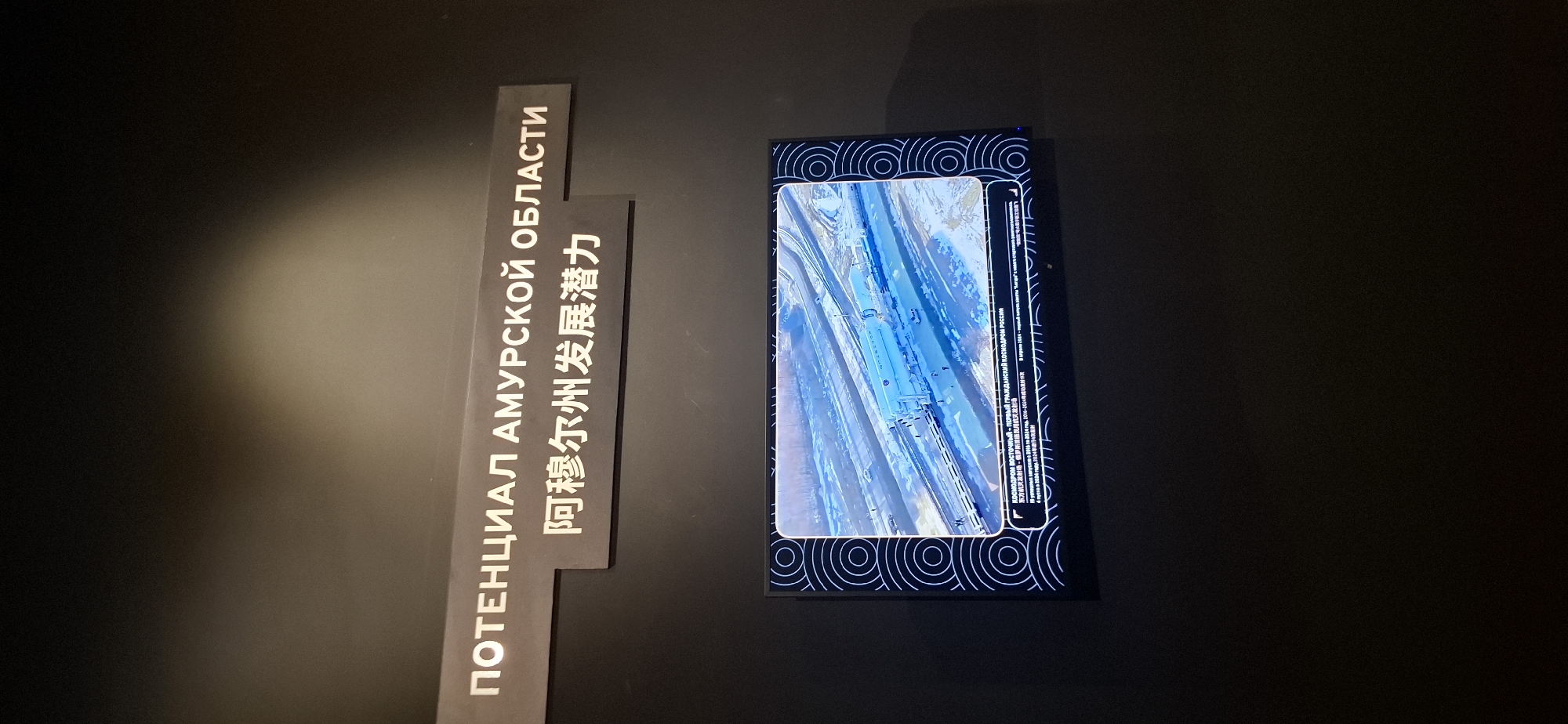 A dark wall displays a horizontal sign with white lettering in Russian and Chinese, reading "POTENTIAL OF AMUR REGION" and "Amur Region Development Potential". Below this, a large, flat-screen television displays an aerial view of a launch vehicle on a railway track, seemingly in a snowy environment. The screen has a decorative border of concentric circles on the left and right edges. Text in Russian appears at the bottom of the screen, identifying the scene as the "Vostochny Cosmodrome - the first civilian cosmodrome of Russia". The lighting in the scene is dim, with a spotlight illuminating the sign from above.