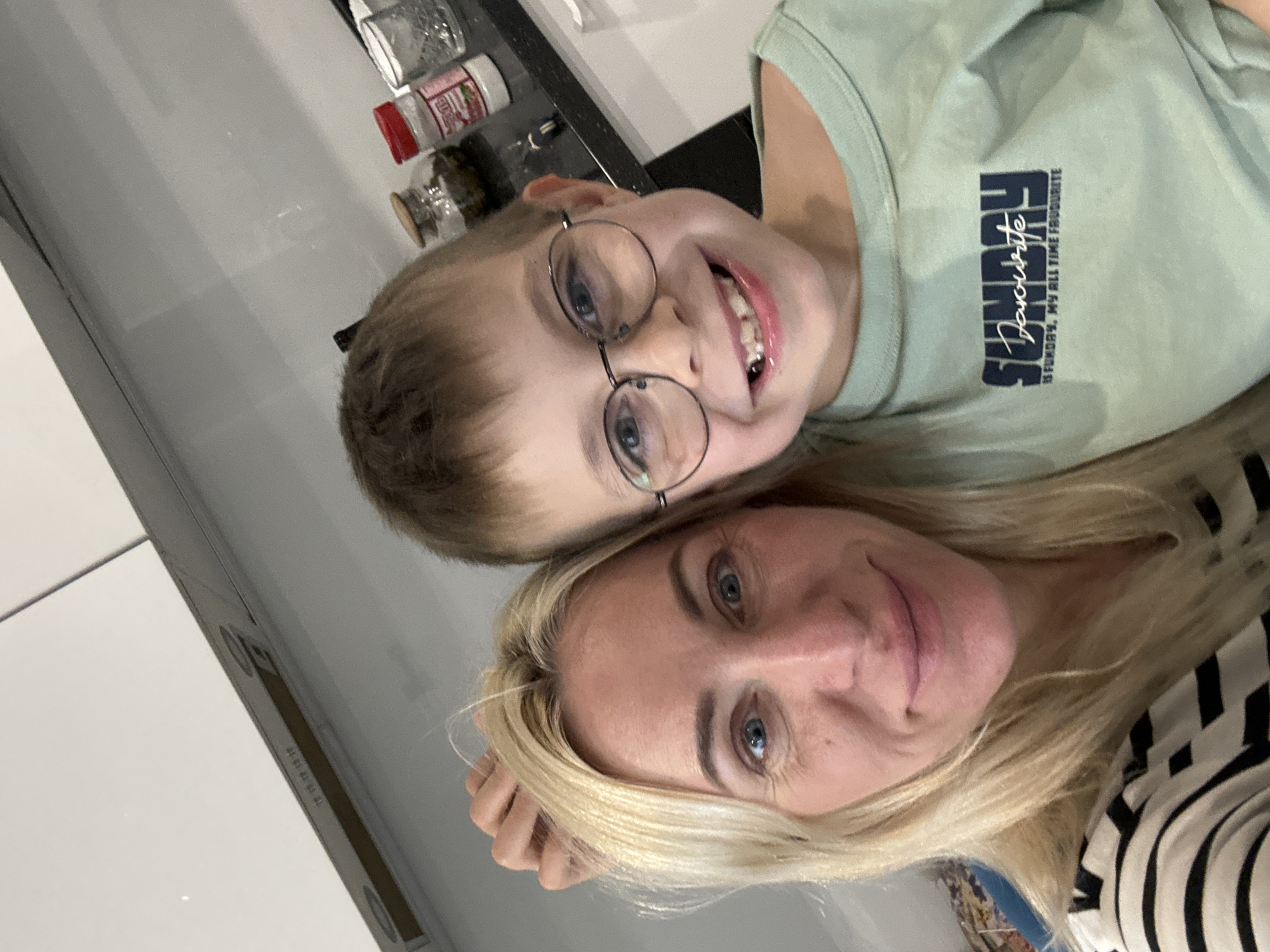 The image shows a selfie taken indoors. A woman with blonde hair, wearing a striped black and white shirt, is looking directly at the camera. Leaning close to her is a young boy with short brown hair and glasses. He is smiling, revealing braces. He is wearing a light green t-shirt with the word "Sunday" printed on it. In the background, there are kitchen items such as jars and containers on a countertop. The location is in a city of Unknown, Unknown.