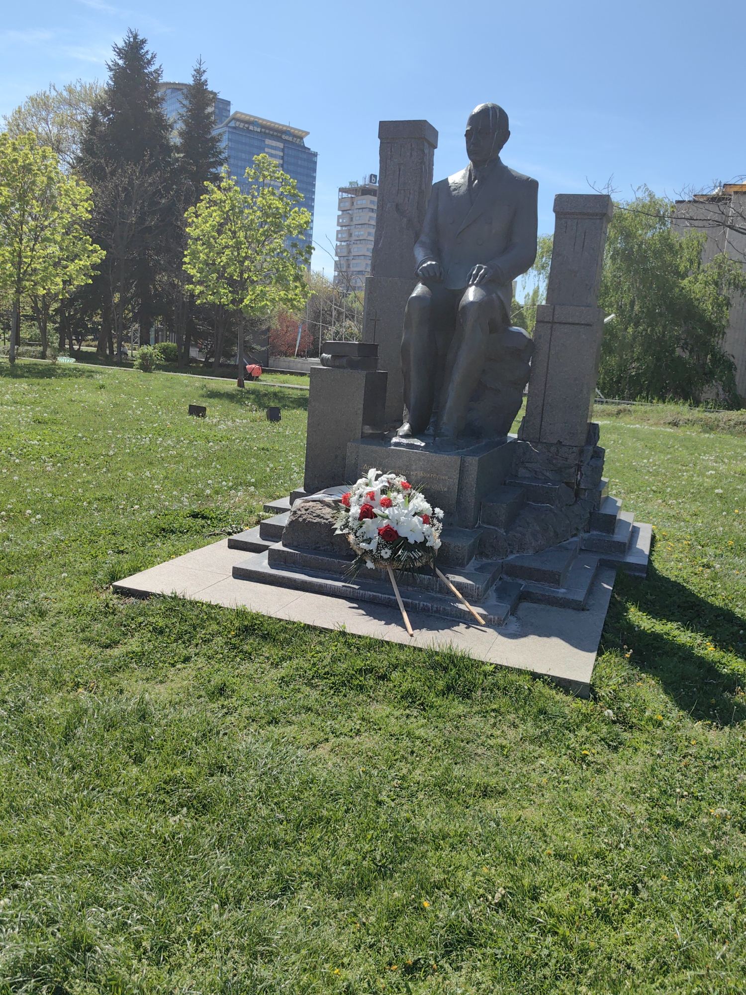 The image depicts a seated statue of a man in a park setting. The statue is made of a dark material, possibly bronze or granite, and is positioned on a multi-tiered stone base. A wreath of white and red flowers is placed in front of the statue.
The background includes trees, a grassy field, and modern buildings in the distance. The sky is clear and blue.
The location is likely in Sofia, Bulgaria.