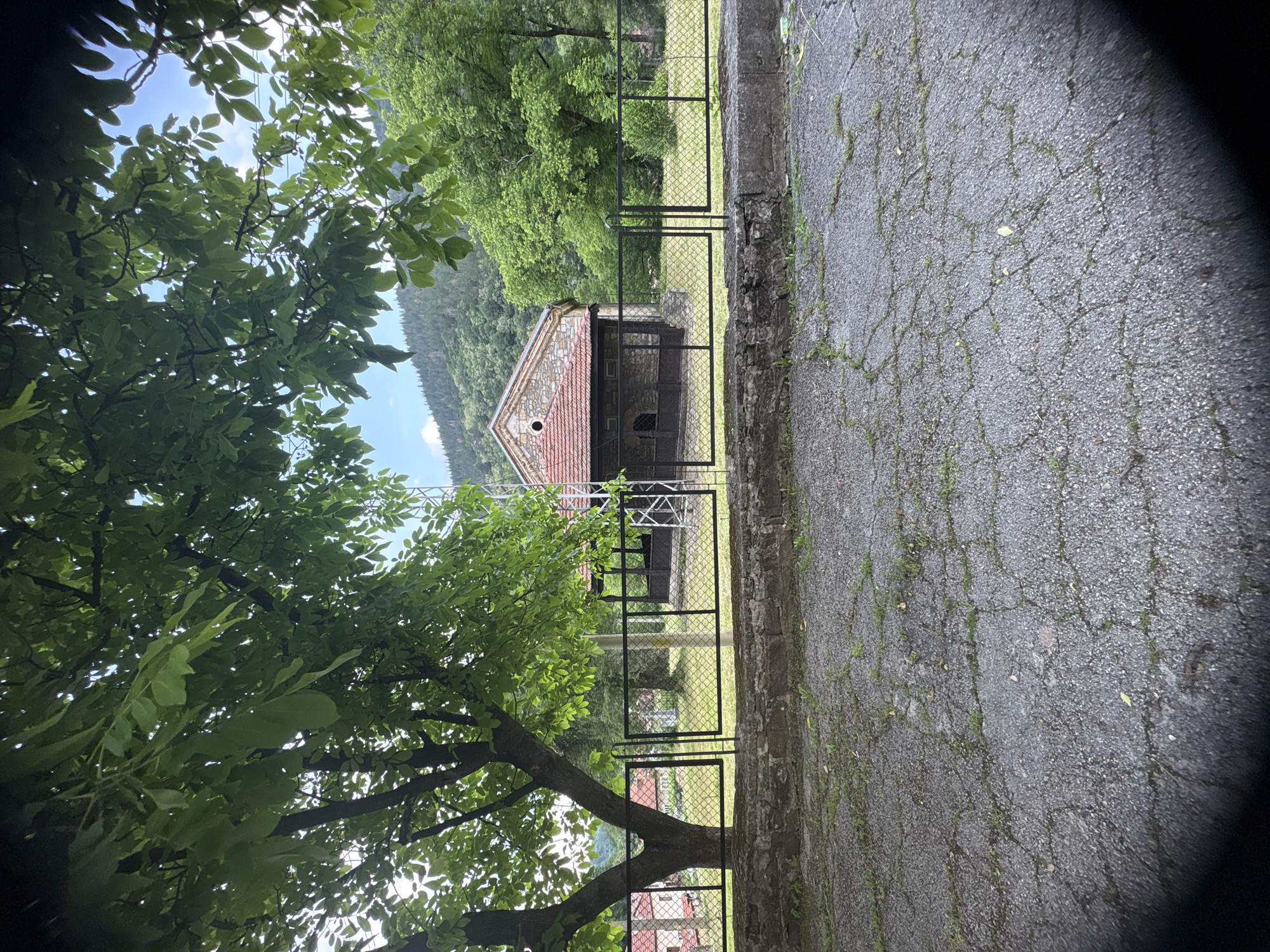 The image depicts a rural or semi-rural outdoor scene in Unknown, Unknown, under bright daylight conditions. The foreground features a weathered, cracked asphalt or paved surface with sparse weeds and moss growing in the fissures. Above this, a low stone wall supports a black metal chain-link fence, which appears to be comprised of two contiguous panels, forming a closed barrier.
Behind the fence, a grassy expanse is visible. To the left, a large deciduous tree with dense green foliage partially obscures the view. Its trunk is dark and substantial. Through gaps in the leaves, parts of red-roofed buildings can be glimpsed further in the background, suggesting a village or cluster of homes.
In the center-right of the midground, beyond the fence and tree, stands an older stone building with a gabled roof covered in reddish-brown tiles. The building's facade features a small circular opening near the apex of the gable and recessed, dark openings below, possibly entrances or windows. Adjacent to the left side of this building (from the viewer's perspective), a white metal lattice structure, resembling a small mast or antenna tower, rises. The area around the building is covered in grass and scattered green trees and shrubs.
The background is dominated by a large, tree-covered mountain or hill under a clear blue sky with some wispy white clouds. Dense green forestation extends across the entire mountainous landscape. No people or animals are discernible within the frame. The overall impression is one of a serene, natural environment with a few man-made structures.