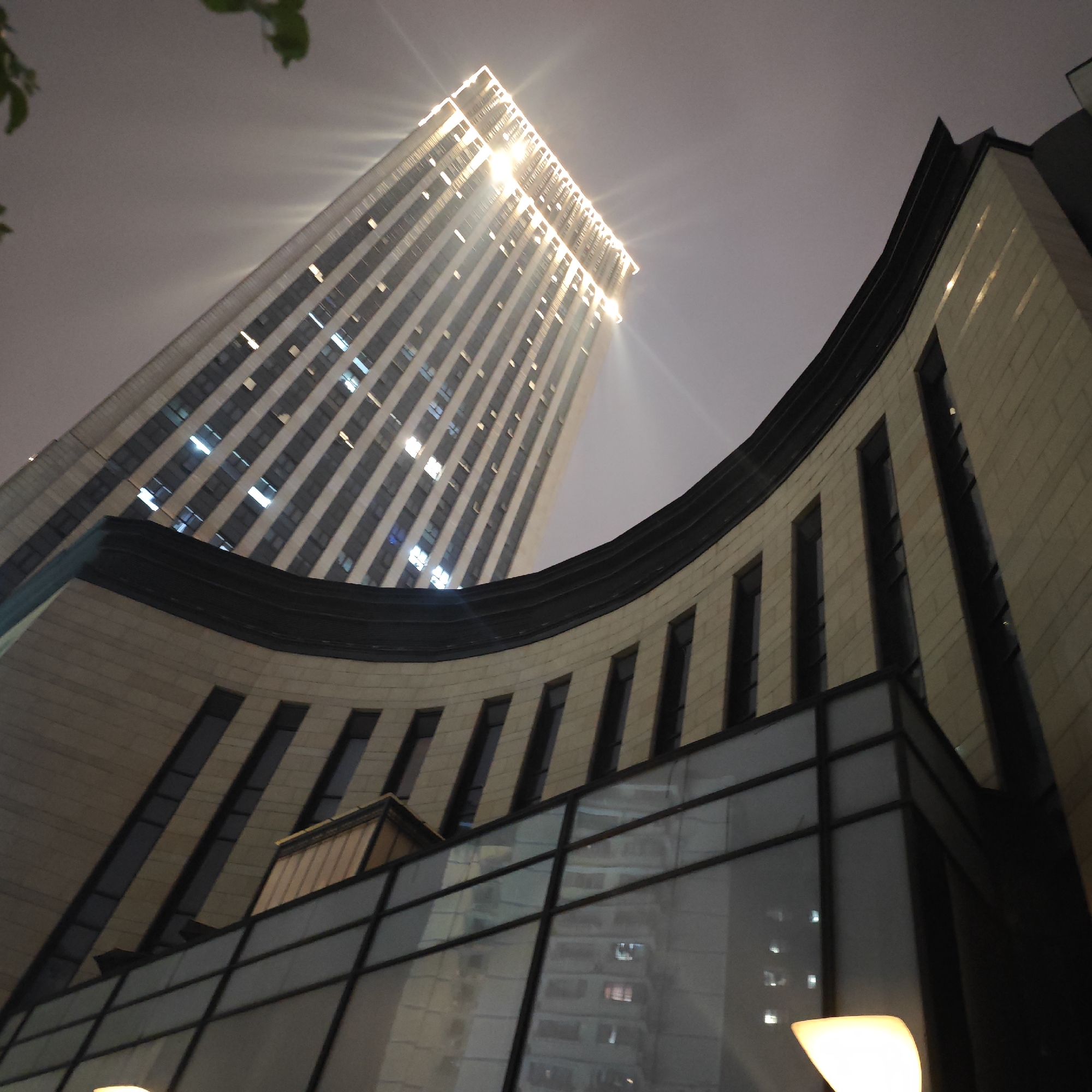The image displays an upward-angled view of two modern buildings at night, likely situated in an urban environment like Yuzhong District, China. In the background, a tall, rectangular skyscraper dominates, its numerous windows and upper edges brilliantly illuminated. The lights on this building create noticeable starburst effects against the dark, overcast sky, suggesting either architectural lighting or interior lights visible through windows.
In the foreground, a distinctively curved building occupies the lower and right side of the frame. Its facade is composed of light-colored, possibly stone or paneling, featuring evenly spaced, narrow, vertical windows. The lower section of this curved building includes large, reflective glass panels, which show faint reflections of interior spaces and possibly other structures. A dark, curved architectural element forms the top edge of this foreground building, separating it from the background sky and the distant skyscraper. A bright, diffused light source, possibly a lamp, is visible in the bottom right corner, casting a warm glow. Faint, dark foliage is present in the upper left corner. The time of day is clearly night or late dusk due to the dark sky and prominent artificial lighting. No specific activity or people are depicted, and no visible text is present. The scene evokes a sense of modern urban nighttime architecture.