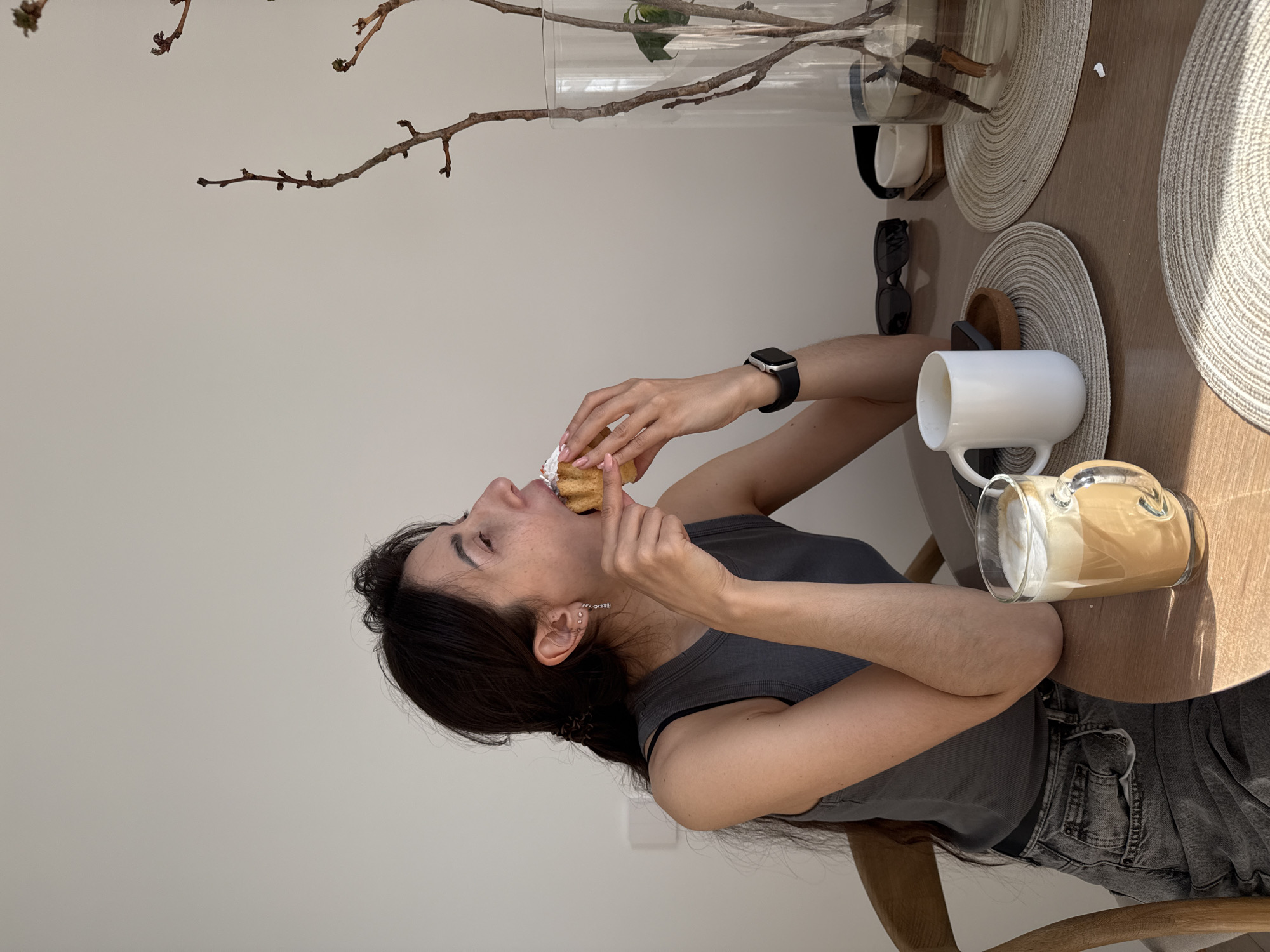 A candid, vertically oriented indoor shot captures a young woman seated at a round wooden table, enjoying a casual meal or snack. The woman, with long dark hair, is dressed in a dark gray tank top and faded gray jeans. She is actively eating a pastry, which appears to be a donut or cream puff generously topped with white cream or frosting, held between her hands as she takes a bite. Her expression indicates focused enjoyment of the food.
On the wooden tabletop, positioned on two light-colored circular placemats, are various items. These include a white ceramic mug, a clear glass mug holding a layered beverage with foam (likely coffee), a pair of dark sunglasses, and a partially visible smartphone. A tall, clear glass vase containing several bare branches or twigs serves as a central decorative element. The background consists of a simple, light-colored wall.
The scene is illuminated by natural daylight, suggesting it is daytime, possibly morning or early afternoon. The overall atmosphere is relaxed and informal. No visible text is present in the image. This interior scene, with its modern minimalist decor, is set in Perm, Russia.