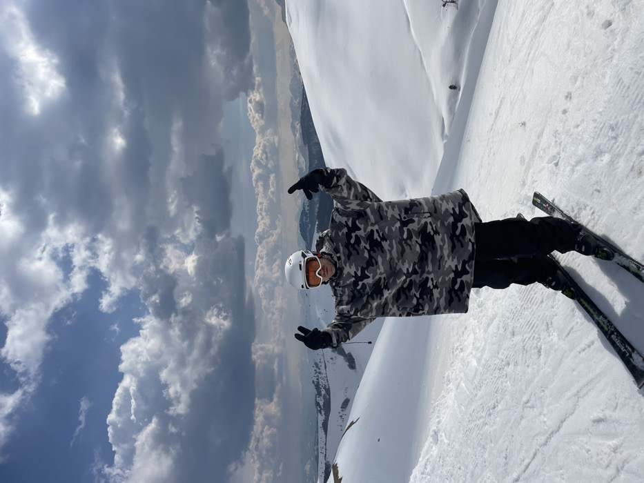 A solitary individual is depicted standing on a snow-covered slope in Katun Bačko brdo, Montenegro. The person wears a white ski helmet with orange-tinted goggles, an oversized grey and black camouflage-patterned fleece poncho, black ski pants, and black gloves. The individual is positioned on green and black skis, with ski boots secured in black bindings. Both hands are raised to chest height, palms facing forward, with the index and middle fingers of each hand extended in a V-shape. The background presents an expansive view of snow-covered mountains and valleys under a sky featuring significant formations of dark grey cumulus clouds interspersed with lighter blue patches.