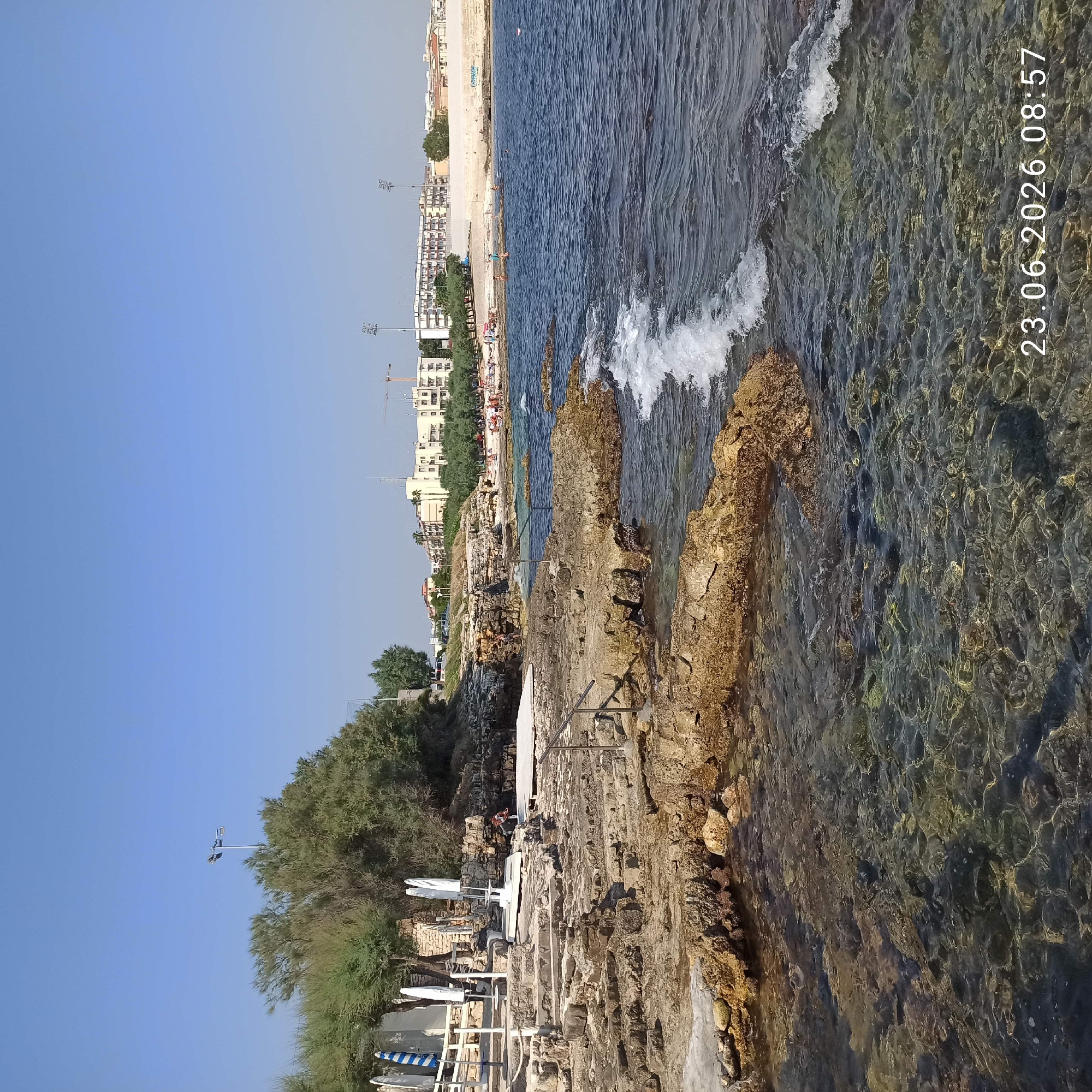 The image depicts a rocky coastline in Molfetta, Italy, on a clear, sunny day. The foreground features the shallow, clear water with visible rocks beneath the surface. The rocky shore is uneven and textured, with some areas appearing to be man-made steps leading down to the water, reinforced with metal railings in some sections. Further back, there are beach umbrellas and seating areas set up on a wooden platform, suggesting a recreational or beach club area. Vegetation, including trees and bushes, grows along the top of the rocky embankment. In the background, a city skyline is visible, characterized by multiple buildings, some of which appear to be apartment complexes or hotels, along with some construction cranes. The sea is a deep blue, with small waves breaking against the rocks. A few people are scattered on the distant sandy beach and in the water, indicating a public beach area. A date and time stamp, "23.06.2026 08:57", is visible in the lower right corner of the image.