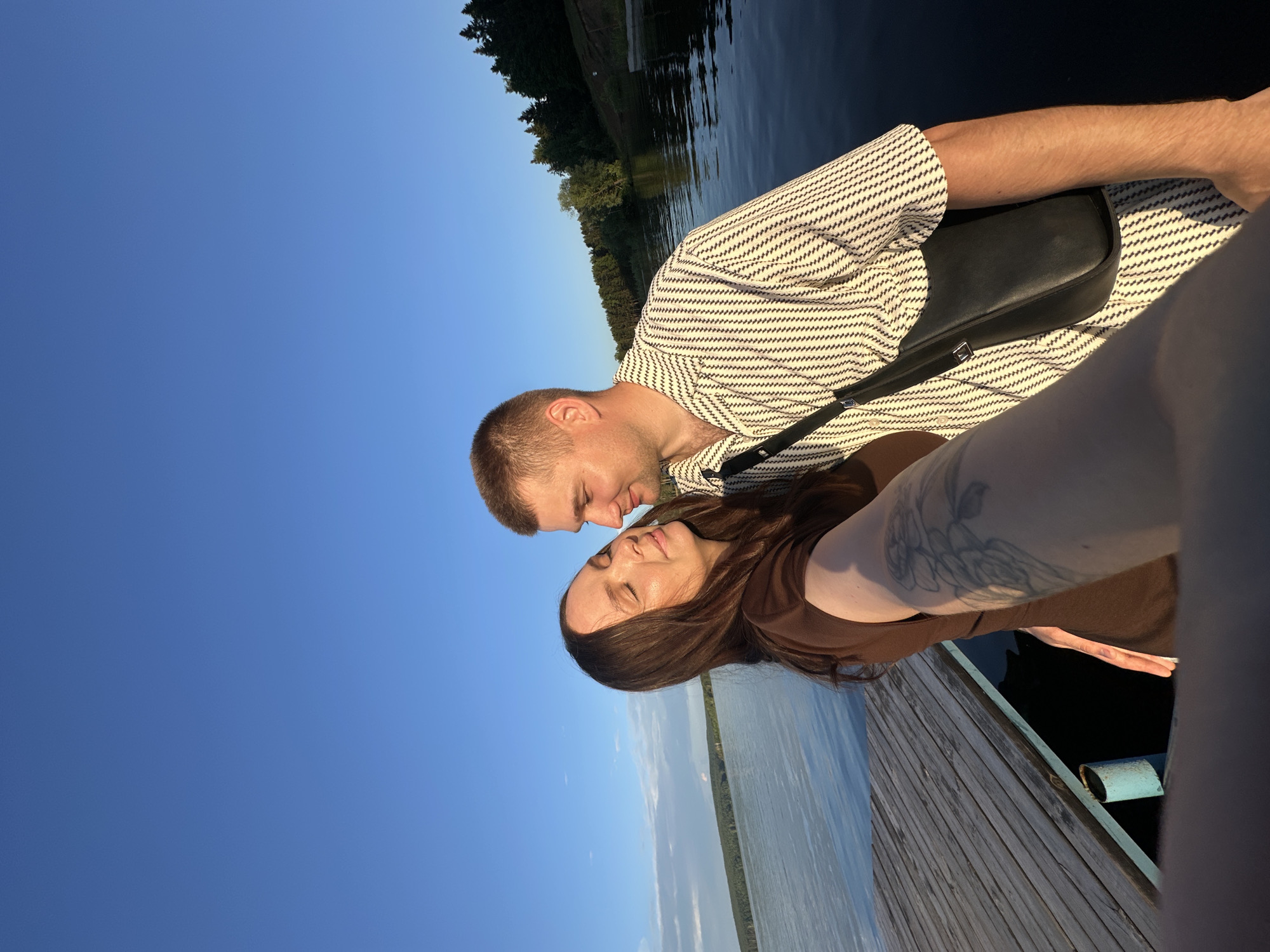 A man and a woman are standing on a wooden dock by a body of water. The man is wearing a striped shirt and a black bag. The woman is wearing a brown dress and has tattoos on her arm. They are looking at each other, with the woman's eyes closed. The background shows a lake with trees and a clear blue sky. The scene appears to be set in the city of Chaikovsky, Russia, as indicated by the presence of a similar dock and body of water often seen in photographs from this location. The lighting suggests it is either sunrise or sunset.