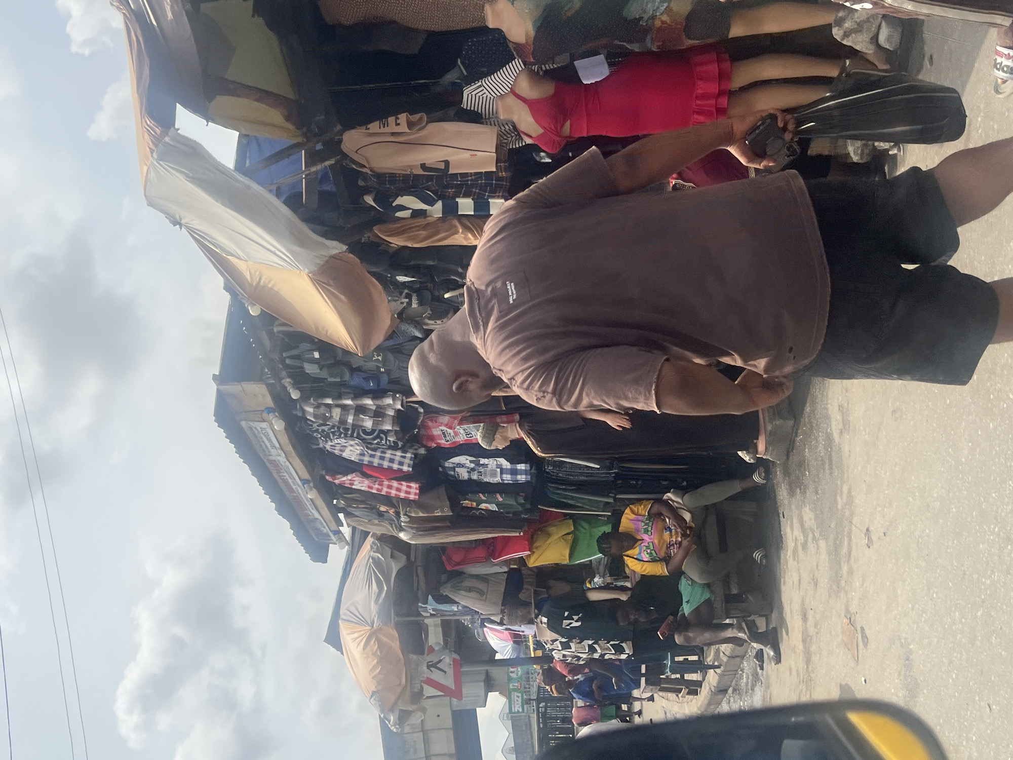 A market scene in Alaiyabiagba, Nigeria, viewed from ground level. Overhanging fabric shelters display assorted clothing. People are browsing. One man is in the foreground. He is bald, wearing a brown T-shirt and black shorts. The sky is overcast. A vehicle is partially visible in the bottom foreground.