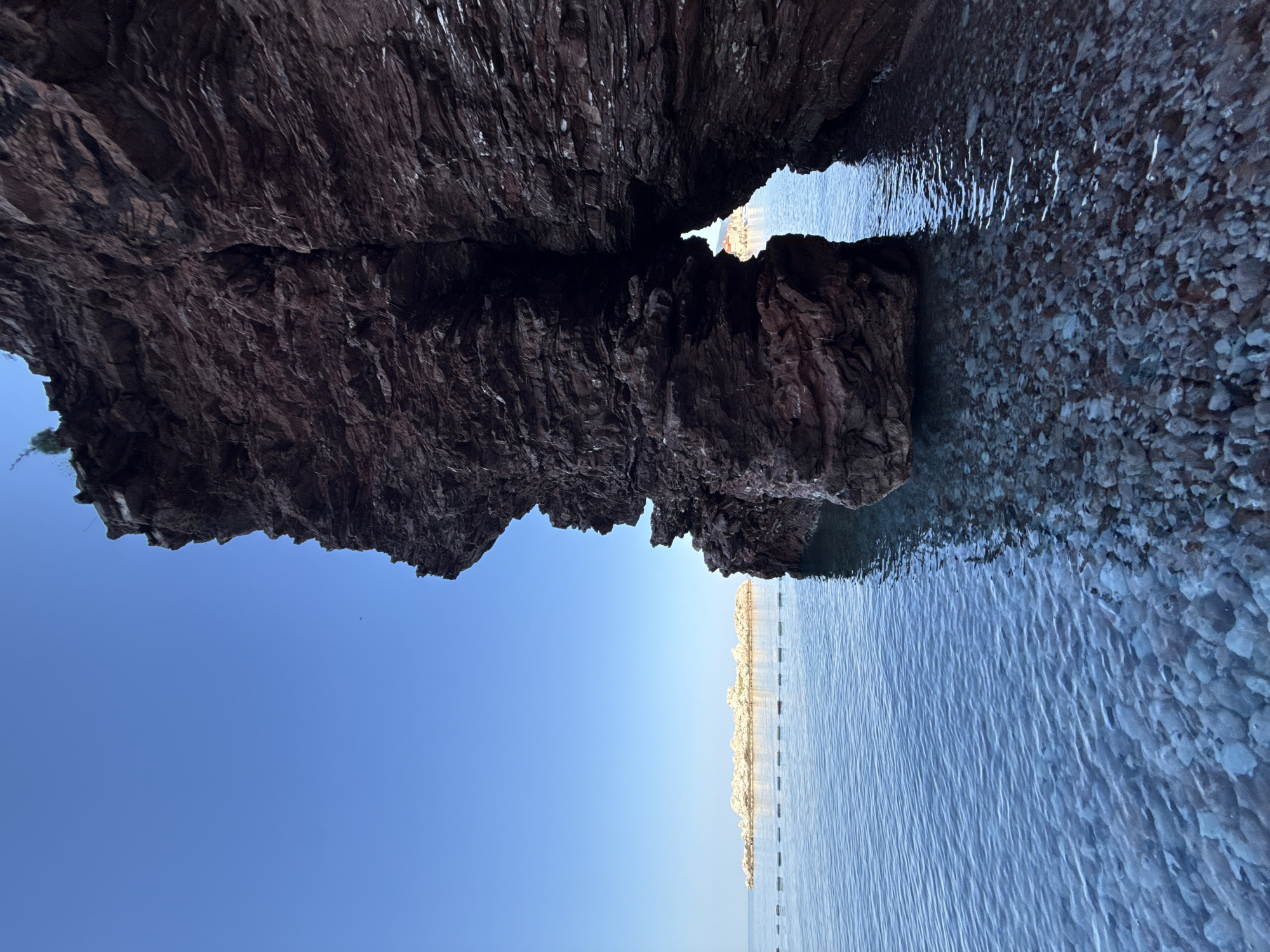A jagged, dark red rock formation dominates the right side of the frame, creating a natural archway. Through the arch, a clear view of the azure sea and a distant, rocky islet can be seen. The foreground consists of shallow, crystal-clear water revealing a seabed covered in smooth, light-colored pebbles. The water is calm, with gentle ripples reflecting the clear blue sky above. To the left, the sea stretches towards the horizon, where the same rocky islet is visible, with a thin line of buoys marking a boundary in the water. The overall scene evokes a sense of natural beauty and tranquility in the coastal area of Sveti Stefan, Montenegro.