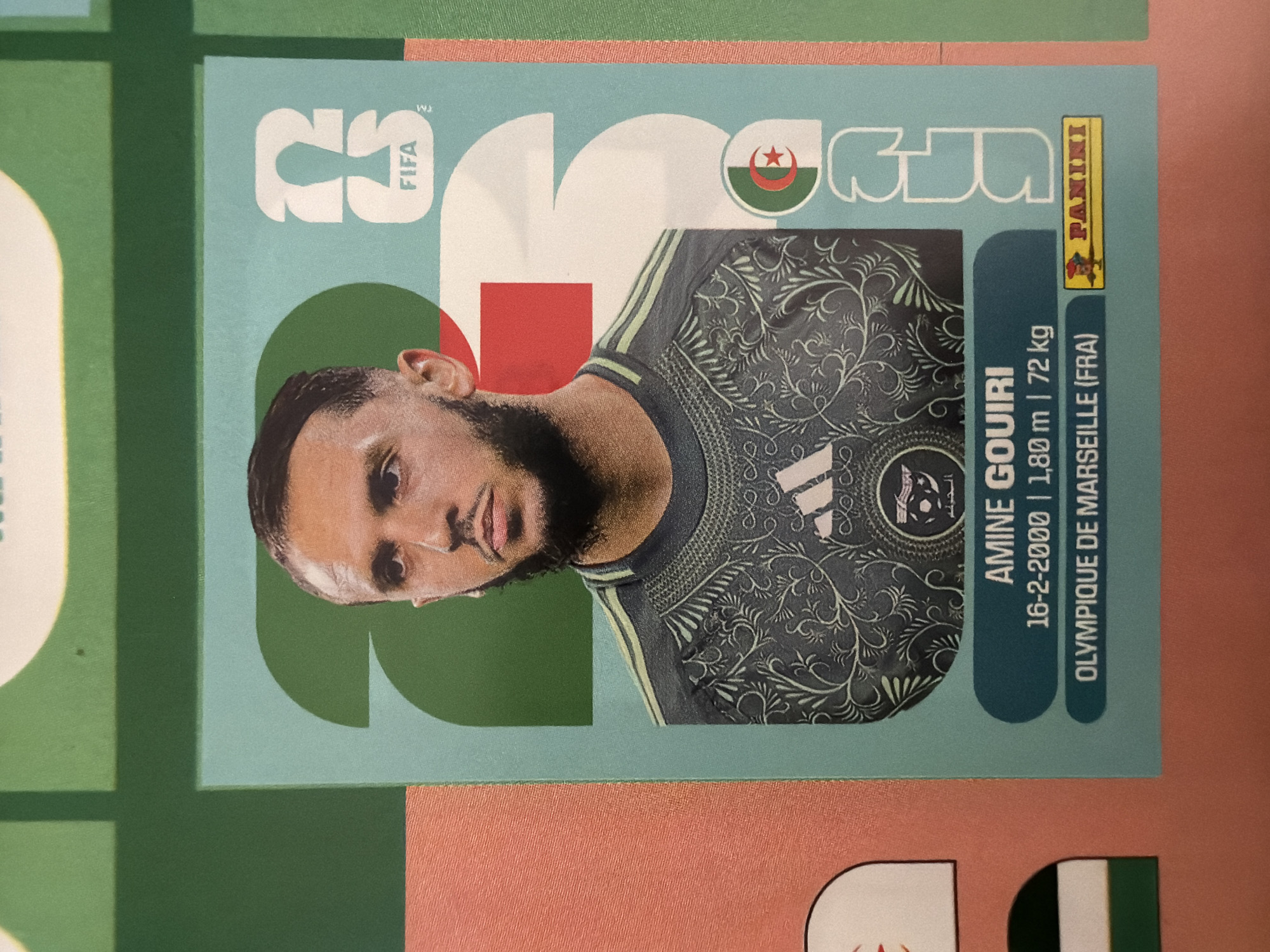 This is a close-up, eye-level, portrait-oriented photograph of a trading card featuring Algerian footballer Amine Gouiri. The card has a light blue background with large, stylized white numbers "2" and "3" overlaid, with the FIFA logo visible near the top right. Gouiri is shown from the chest up, wearing a dark jersey with a green and yellow striped collar and a black and green patterned design on the body, which includes the Adidas logo and a team crest. The Algerian flag is subtly incorporated into the card design, with a red square and a green and white section featuring a red star and crescent visible to the right of Gouiri's head. Below the image of Gouiri, text indicates his name, birthdate (16-2-2000), height (1.80m), weight (72kg), and club (Olympique de Marseille (FRA)). A Panini logo is also present at the bottom right. The card is placed on a surface with pink and green elements visible in the background, suggesting it's part of a larger collection or display, possibly in Zlatitrap, Bulgaria, though the image provides no direct visual evidence to confirm this location.