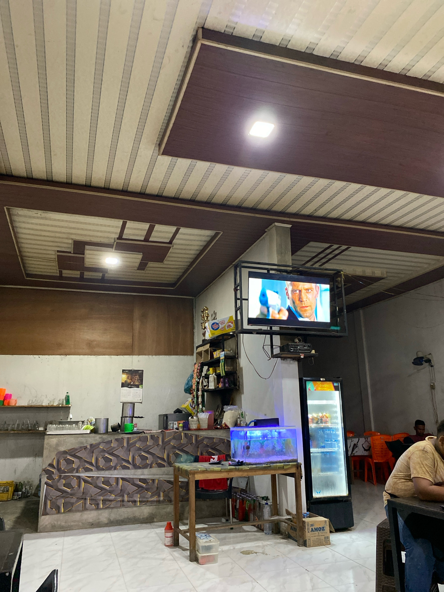 The image depicts the interior of a casual eatery or restaurant in Lhokseumawe, Indonesia. The ceiling is constructed with white and brown panels, featuring a central decorative recessed area with lighting. A flat-screen television mounted on a black metal frame displays a man holding a gun, suggesting an action movie is playing. Below the television, a small electronic device is visible.
The scene includes a bar area with shelves stocked with various items, including bottles and what appear to be food products. A small aquarium with blue lighting is placed on a wooden table. Adjacent to the aquarium is a tall, illuminated commercial refrigerator filled with drinks. On the right side of the image, there are orange plastic chairs and tables, with a few patrons seated in the background, one of whom is visible wearing a yellow shirt. A patterned wallpaper covers a section of the wall behind the bar. A calendar hangs on the wall above the bar.
The floor is tiled with a light-colored pattern. Various objects are scattered around, including bottles, containers, and a wooden chair with a table. The overall impression is of a modestly furnished establishment catering to a local clientele.