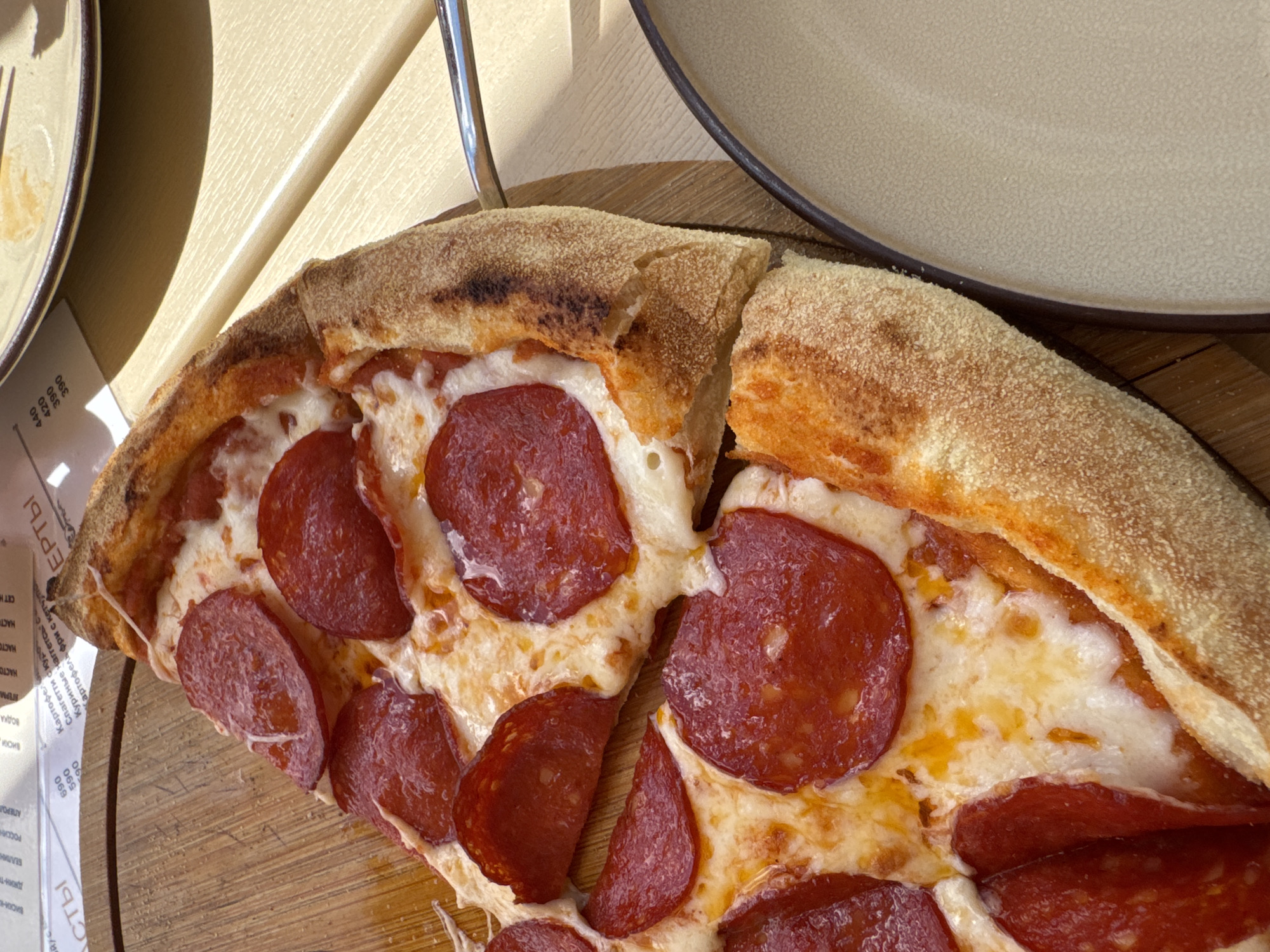 A close-up, top-down view of a pepperoni pizza slice on a wooden board is presented, with a portion of a beige plate and a metal serving utensil visible in the foreground and right side of the image, respectively. The pizza slice features a thick, golden-brown crust generously topped with melted mozzarella cheese and numerous slices of pepperoni. The background includes a blurred menu with Cyrillic text and prices, suggesting a restaurant setting. The scene is illuminated by natural light, casting soft shadows. The location is identified as Abrau-Dyurso, Russia.