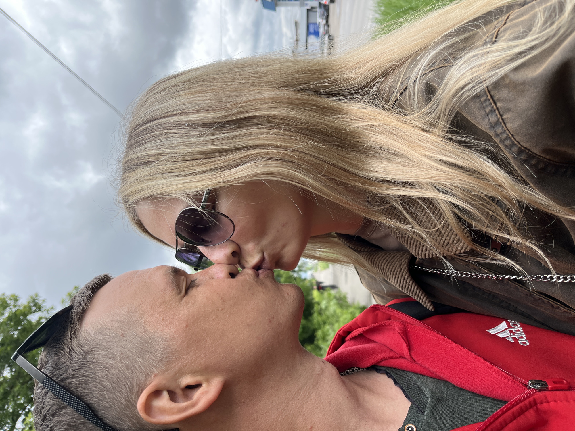 A close-up, eye-level shot depicts a woman and a man kissing. The woman has long blonde hair, sunglasses, and is wearing a brown jacket with a chain detail. The man has short hair and is wearing a red Adidas jacket. The background shows a cloudy sky, trees, and a street with buildings. Based on available information, this scene is located in the city of Chaikovsky, Russia.