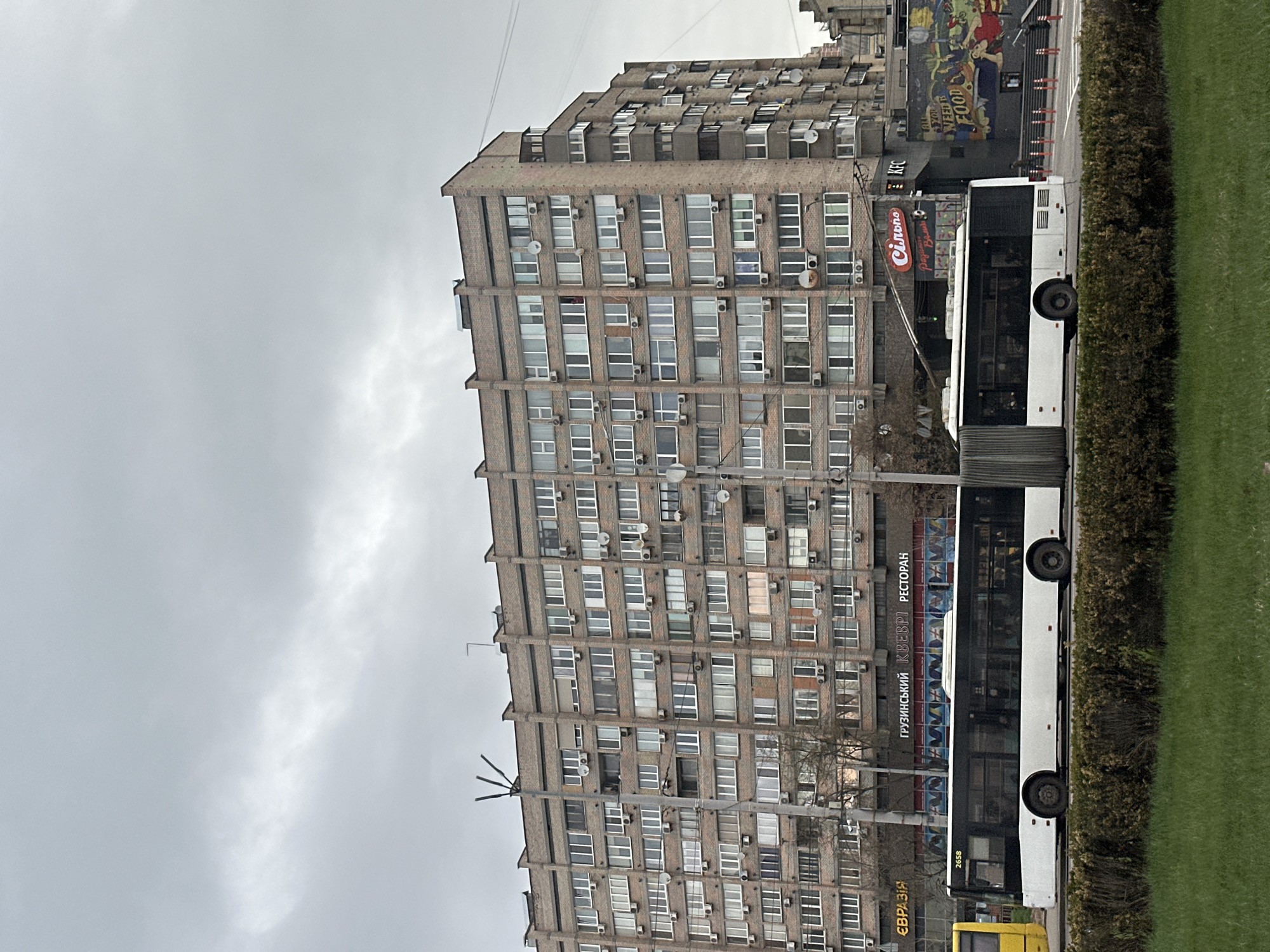 The photograph shows a multi-story building in Kyiv, Ukraine. The building is constructed of brick and has numerous windows, many of which have air conditioning units. A white public bus is parked along the street in front of the building, partially obscuring a yellow taxi. The bus advertises a Georgian restaurant. Vegetation, including grass and bushes, occupies the foreground. The sky is overcast. A building with a large mural can be seen next to the grey apartment building. A KFC restaurant appears to be present.