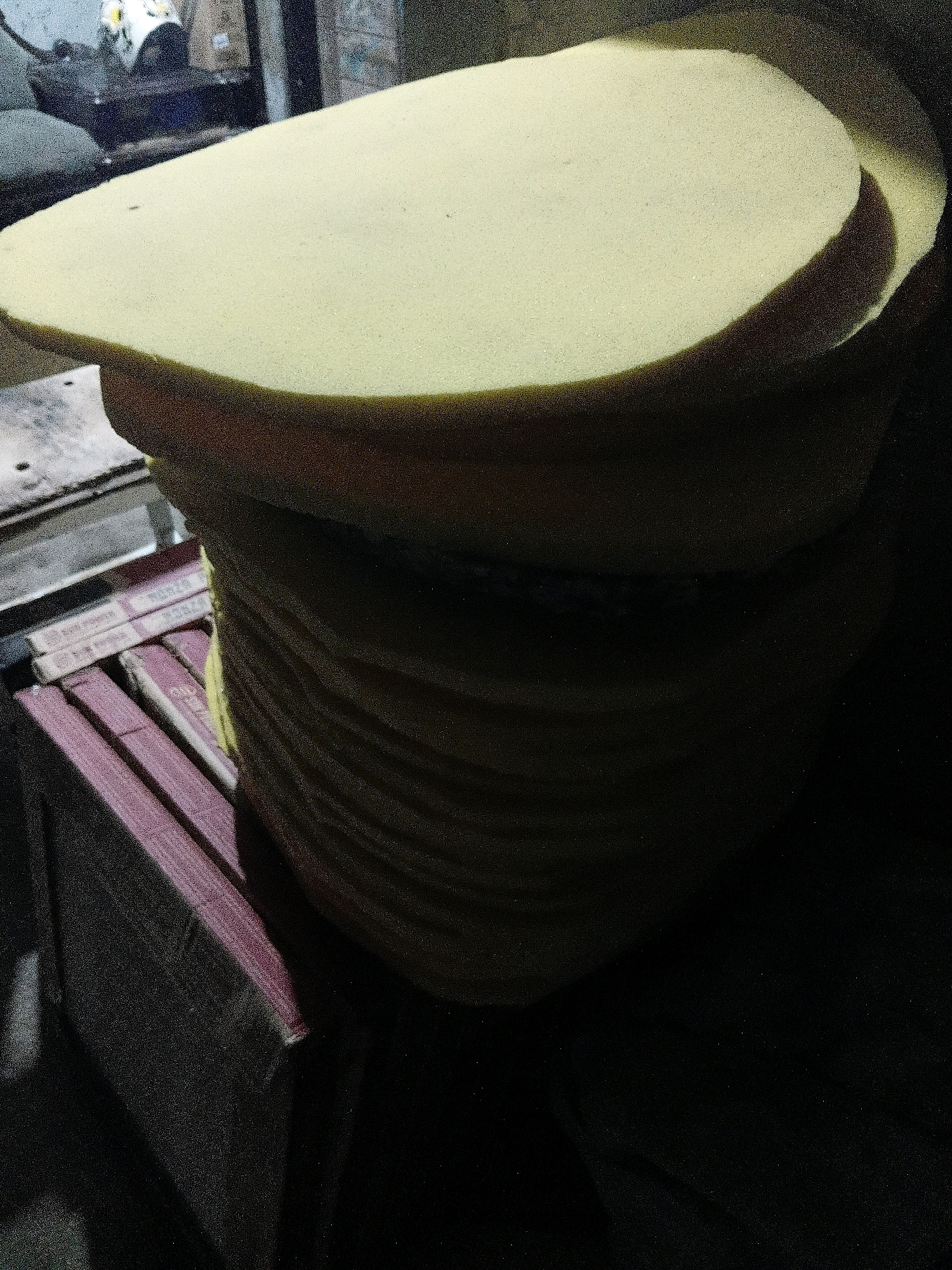 In Cikal, Indonesia, a stack of round, yellow foam cushions is displayed on a table. The cushions are of varying sizes and are neatly piled on top of each other. In the background, there are some objects that are unclear due to the low lighting and focus. The scene is indoors, likely a workshop or a retail space, with some tools and materials visible on the table. The lighting is dim, creating shadows that add to the mysterious atmosphere of the scene.