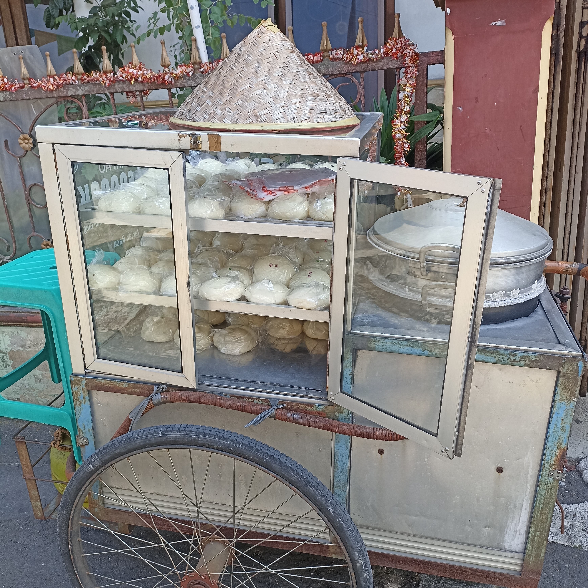 This is a street-level medium shot, captured outdoors in the city of Magelang, Indonesia. The scene features a mobile food cart laden with steamed buns. The cart, constructed of metal with a glass display case, is weathered and shows signs of rust. Inside the display case, numerous white, bun-shaped food items, individually wrapped in plastic, are neatly arranged on shelves. A large, conical woven bamboo lid, typically used for steaming, sits atop the display case. To the left of the cart, a bright turquoise plastic stool is visible. Behind the cart, a rusty metal fence is decorated with festive red and gold tinsel, and greenery can be seen in the background. A large, silver steaming pot is partially visible to the right of the display case, placed on a metal surface attached to the cart. The cart is equipped with a single wheel at the front, also showing signs of wear. The ground is paved with asphalt and stone. No people are present in the frame.