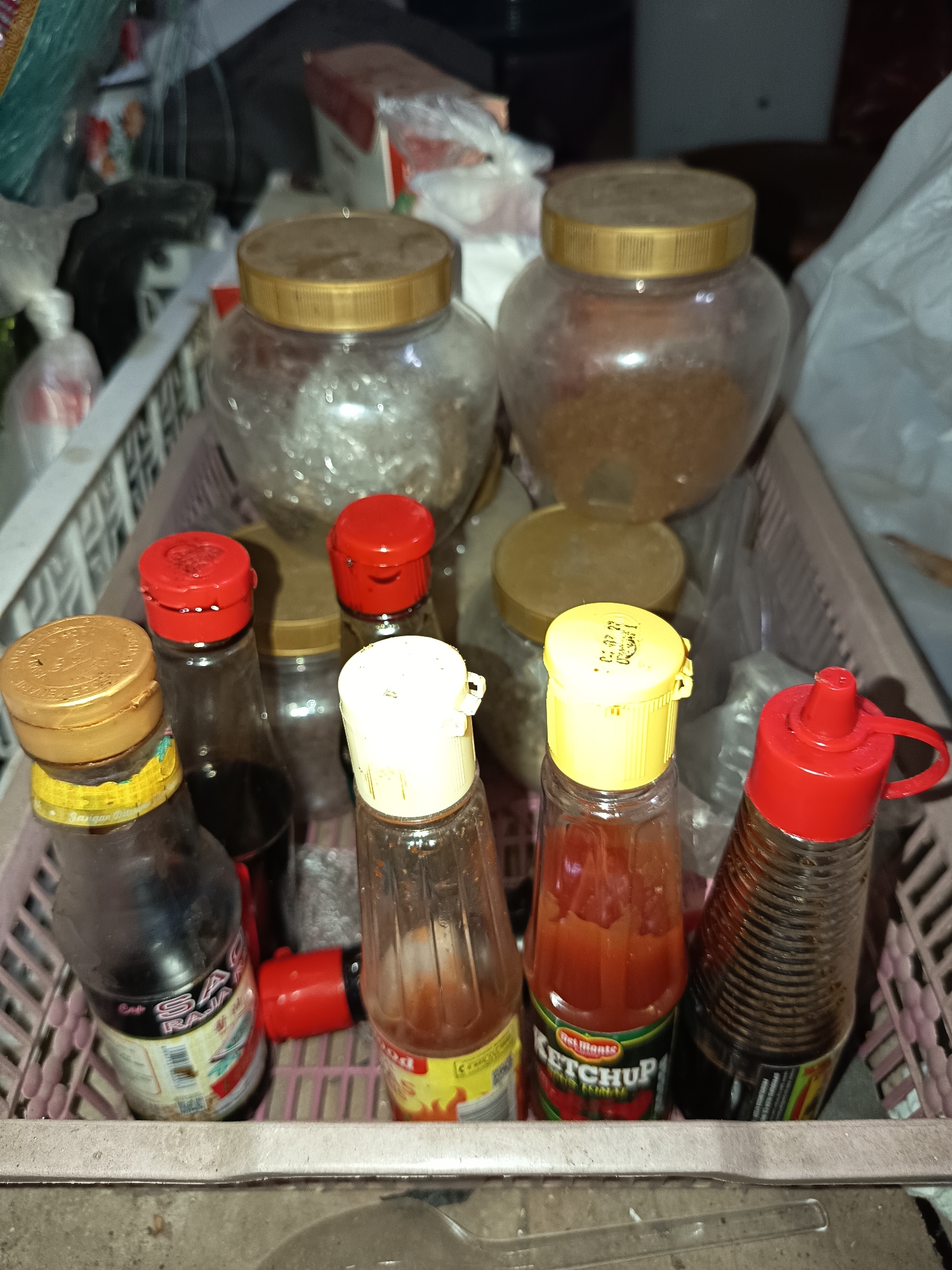 In Krampilan, Indonesia, a basket contains various condiment bottles. Two large glass jars with gold lids are visible in the background, one filled with what appears to be granulated sugar or spices, and the other with a clear, granular substance. In the foreground, a pink plastic basket holds several bottles. From left to right, there is a dark bottle with a gold lid, a bottle with a red lid, a bottle with a yellow lid, a bottle with a red lid, and a dark ribbed bottle with a red flip-top cap. One bottle is labeled "Del Monte KETCHUP." The lighting is dim, suggesting an indoor environment with artificial light.