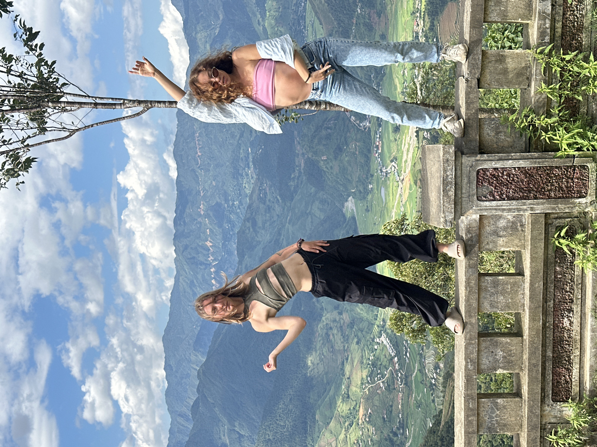 The scene depicts two young women posing atop a weathered stone balustrade overlooking a mountainous landscape in Tu Le, Vietnam. The background features expansive green mountains, deep valleys containing terraced fields, and scattered small villages under a bright blue sky with scattered cumulus clouds.
The woman on the left, with straight, light-colored hair, stands in a wide stance on the balustrade. She is wearing a dark olive green sleeveless crop top with multiple horizontal cut-outs across the chest and midriff, and loose-fitting black cargo pants. Her left arm is bent at the elbow, with her hand held upward, palm facing inward. Her right hand rests near her hip. She wears light-toned, open-toed slide sandals.
The woman on the right, with curly, reddish-brown hair and small, oval-framed sunglasses, also stands in a wide stance on the balustrade. She is wearing a strapless pink bandeau crop top and light-wash blue jeans. A light blue, long-sleeved shirt is draped over her shoulders, with the sleeves hanging down her sides. Her left arm is raised above her head, with her hand slightly cupped. Her right hand is positioned near her right hip. She wears light-colored athletic shoes.
A slender, sparsely leafed tree trunk is visible directly behind the woman on the right, extending upwards from below the balustrade. The stone balustrade in the foreground is grey and appears aged, with some dark reddish-brown textured panels incorporated into its design and green foliage growing at its base.