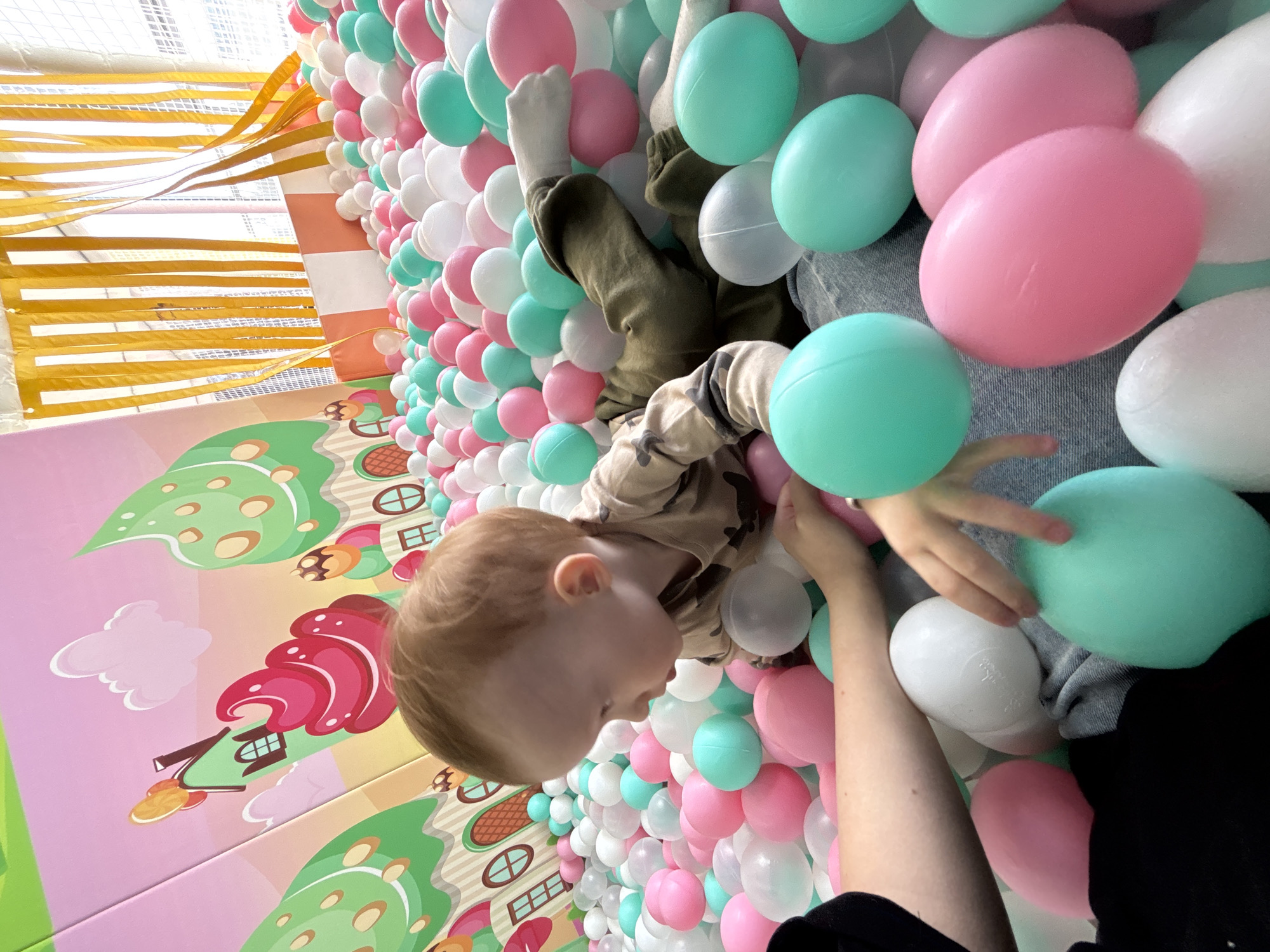 A young child lies in a ball pit in Perm, Russia, surrounded by pink, white, and light blue balls. The pit is decorated with cartoonish images of houses and trees on the surrounding walls. The child is wearing a camouflage shirt. A person's arm is visible, seemingly interacting with the child. Above the pit, there are yellow streamers.