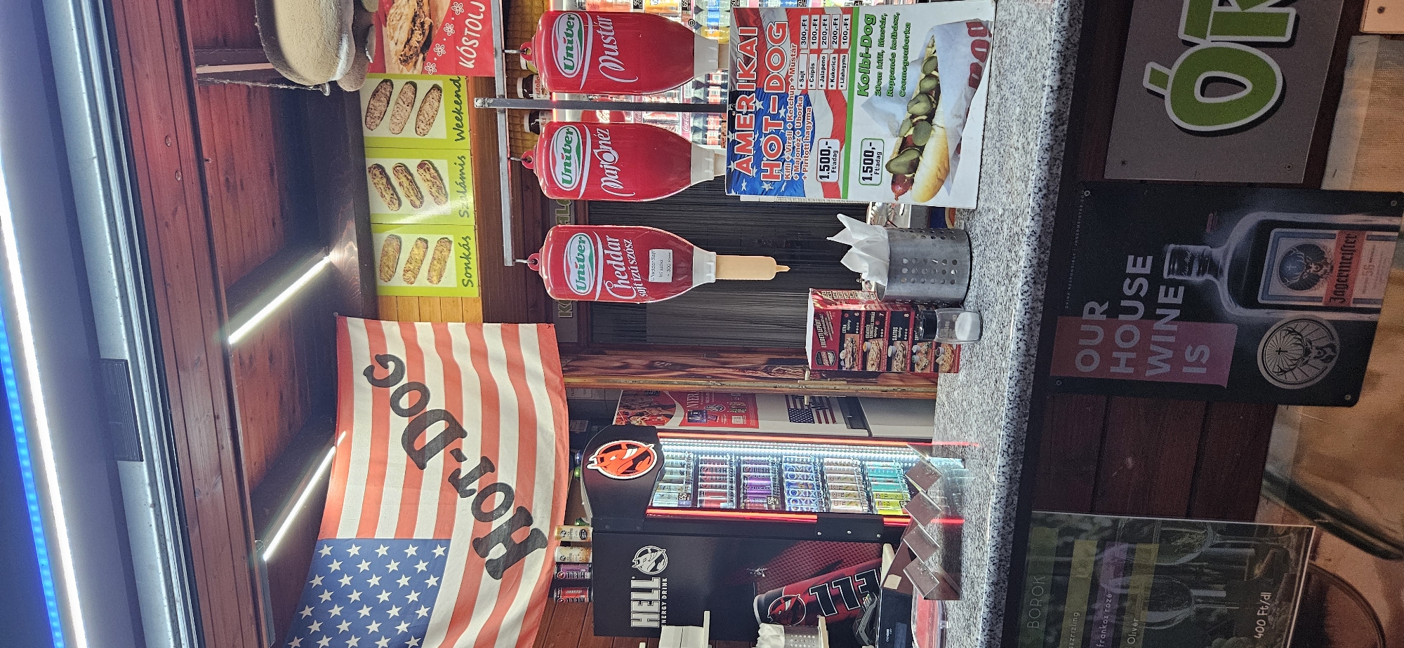 This image depicts an outdoor food stall, likely a hot dog stand, situated in Keszthely, Hungary. The stall is decorated with an American flag emblazoned with "Hot-Dog." Several promotional materials for hot dogs and toppings are visible. Three large red dispensers, likely for condiments like ketchup and mustard, hang from above. A refrigerated display case filled with colorful beverage cans, branded "HELL ENERGY DRINK," is positioned to the left. The counter area displays napkins, a salt shaker, and various menus. In the background, a sign advertising "OUR HOUSE WINE IS JÄGERMEISTER" is prominently displayed. The overall scene suggests a casual, fast-food establishment with a distinctive American theme.