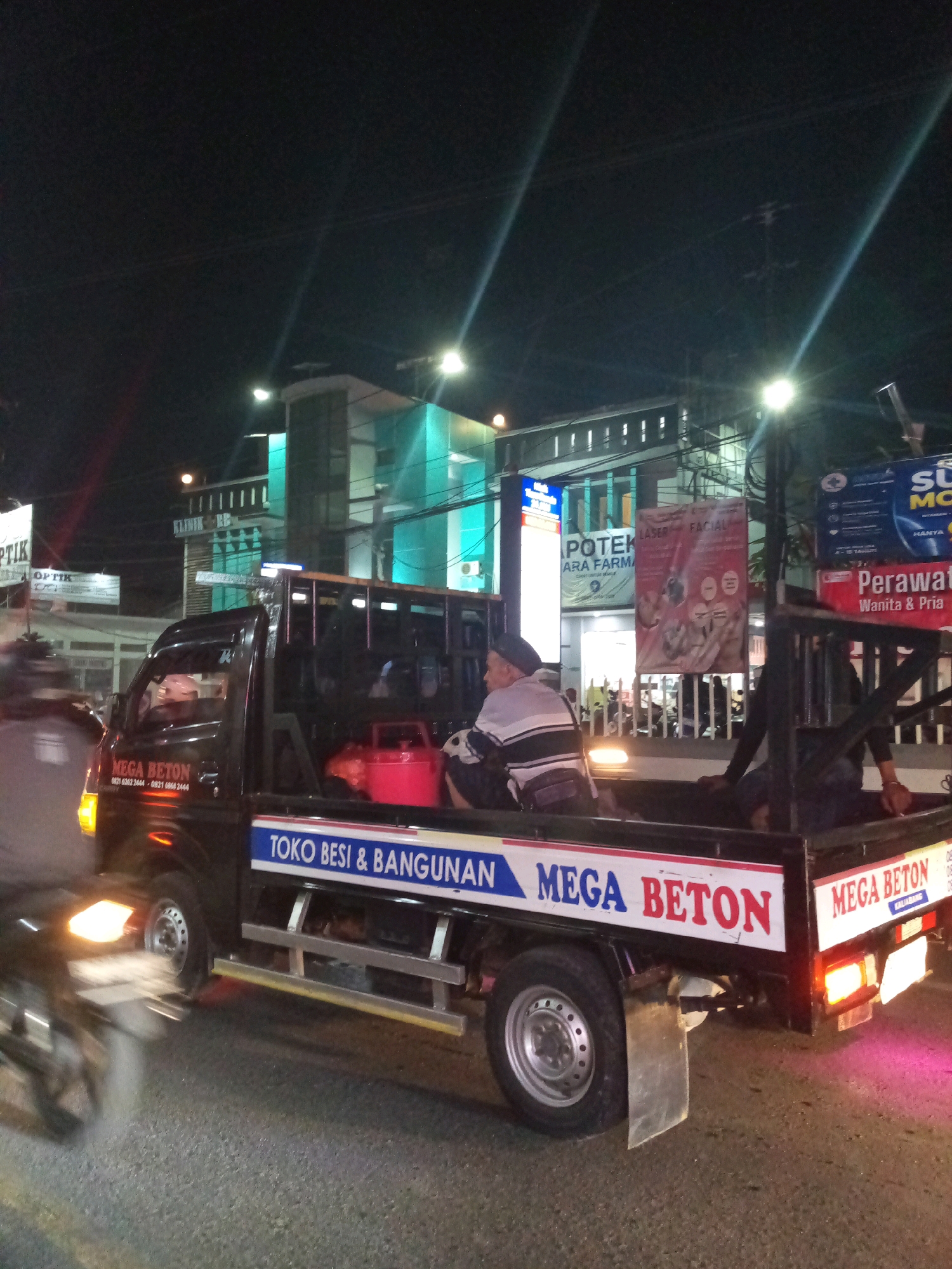 A black pickup truck is parked on a street at night in Bekasi, Indonesia. The truck has text on its side that reads "MEGA BETON" and "TOKO BESI & BANGUNAN". There are two men in the truck bed, one wearing a striped shirt and a cap, and the other partially visible. A red gas tank is also in the truck bed. In the background, there are buildings with illuminated signs, including one that says "KLINIK BB". The street is paved, and there are streetlights visible. A motorcycle is blurred in motion in the foreground.